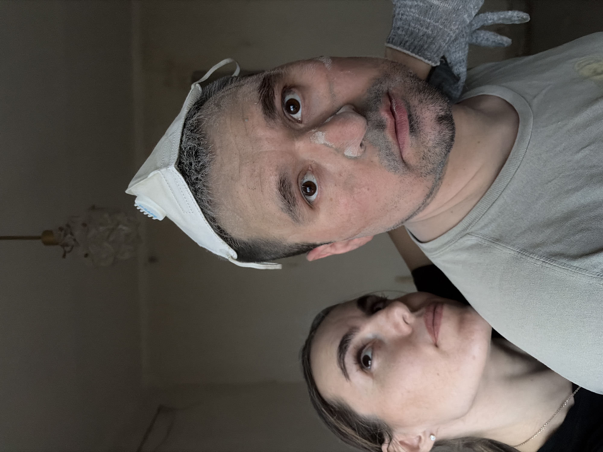 Two people are in an interior. The man in the foreground has a white surgical mask pulled up onto his forehead. He has short dark hair with some gray, and a beard. There is dust on his face. He is wearing a light colored t-shirt and a gray glove. Behind him and slightly to the right is a woman looking up at the camera. She has dark hair. A chain and pendant are visible around her neck. The walls are a light color, and a light fixture is partially visible in the upper left corner. The location is unknown, but is believed to be in Izhevsk, Russia.