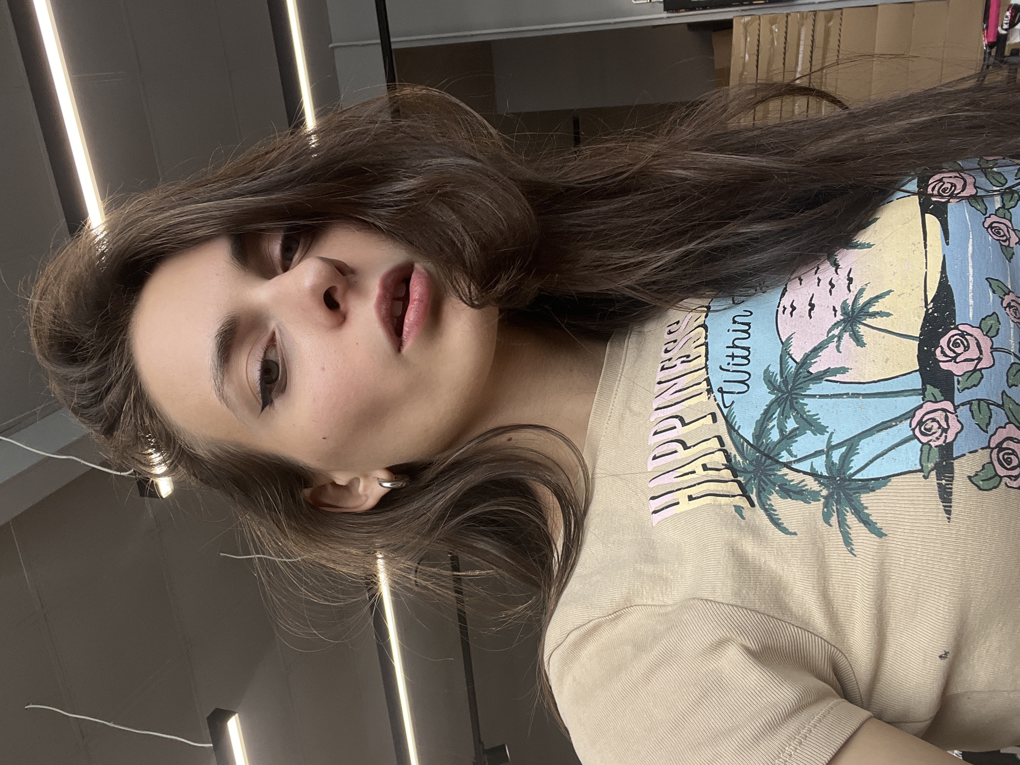 A woman with long, brown, wavy hair is positioned looking down at the camera. She has fair skin, dark eyes, and is wearing minimal makeup, including eyeliner. A silver hoop earring is visible in her left ear. She is wearing a beige t-shirt with a graphic design featuring palm trees, roses, and the word "Happiness" vertically printed.
The background is a commercial or industrial setting, with fluorescent lights visible on the ceiling. Boxes are stacked in the background. The specific location in the city of Krasnoyarsk, Russia cannot be determined.