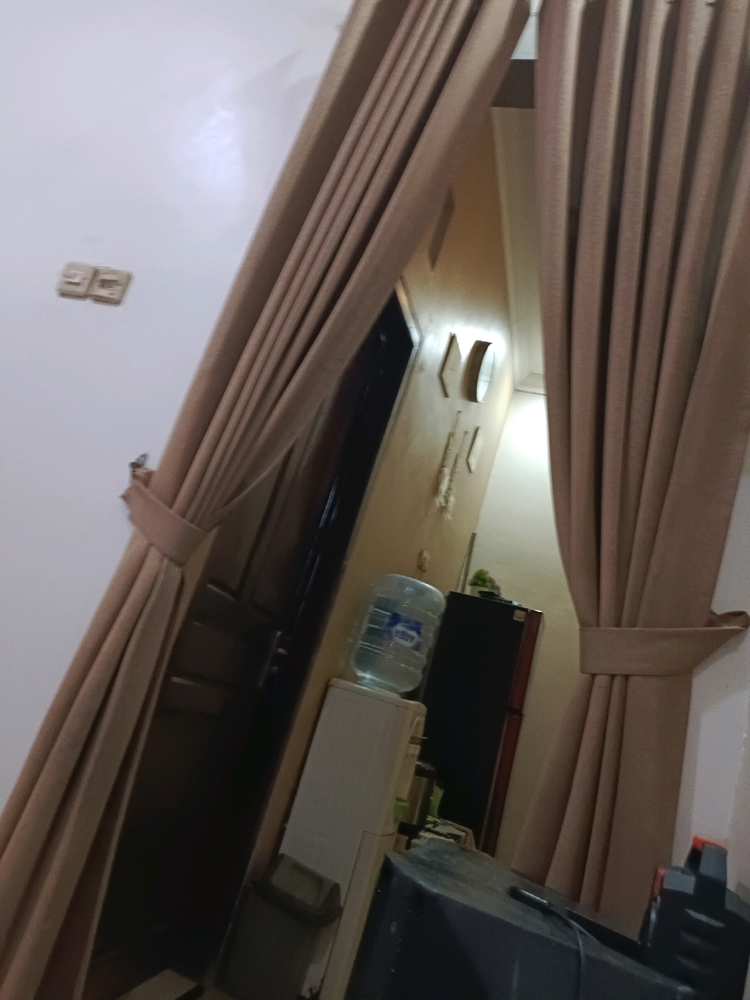 The image depicts an interior scene, likely a residential dwelling, in Rancang Kencono, Indonesia. The foreground is dominated by large, beige curtains that are drawn aside, revealing a hallway or interior space. On the left wall, near the top, are two light switches. Further down the hallway, a dark door is visible. To the right of the door, a large jug of water, possibly from a dispenser, sits on top of a white appliance, which could be a water dispenser or a small refrigerator. Beside the white appliance, a dark upright appliance, likely a refrigerator, is partially visible. On the right side of the image, the beige curtains continue, with a tie-back securing them. In the lower right quadrant, the top of a dark electronic device, possibly a television or a computer monitor, is visible. The lighting suggests it is indoors, with artificial light sources present. The overall impression is a functional, somewhat cluttered living space.