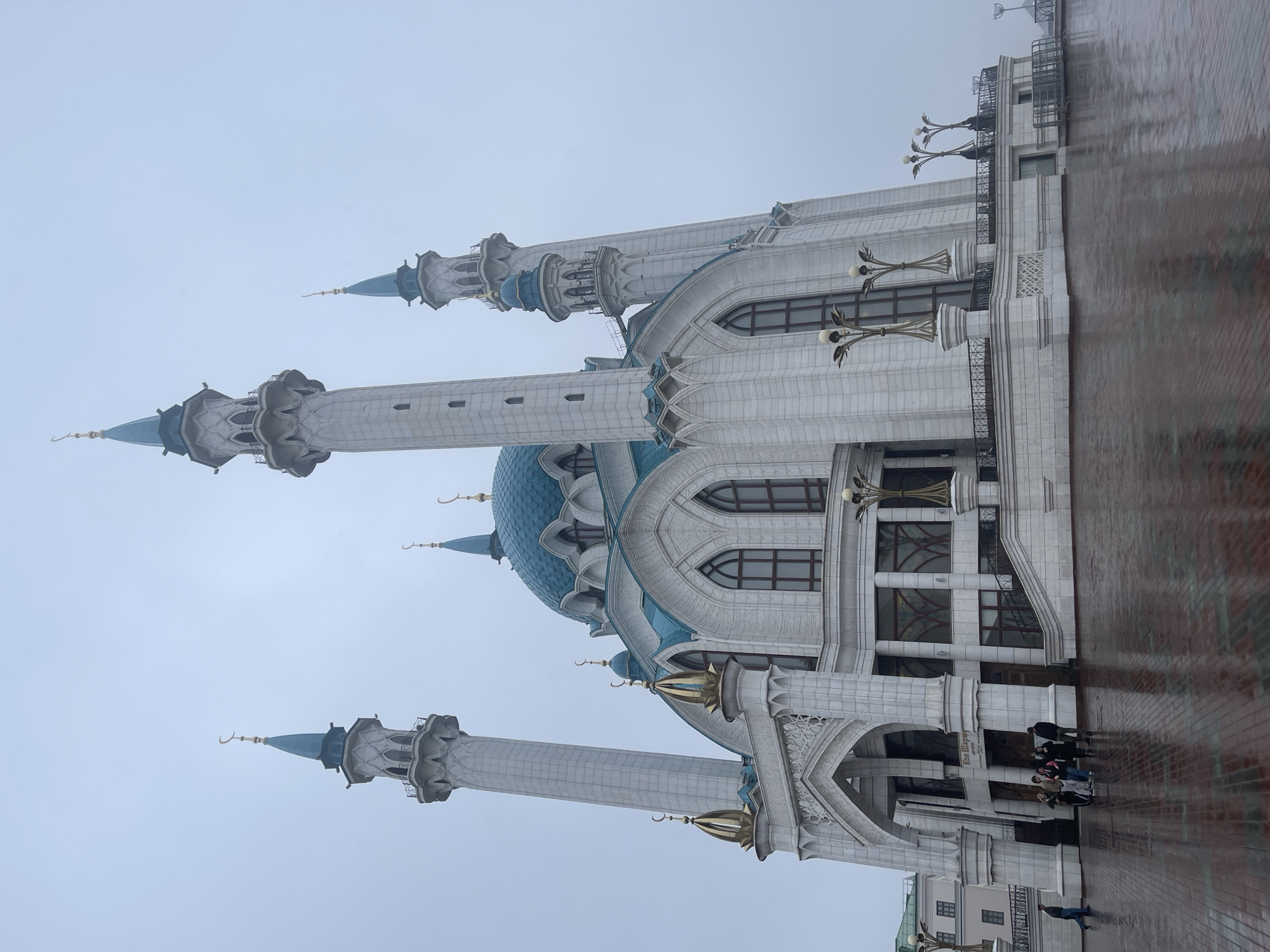The image depicts the Kul Sharif Mosque in Kazan, Russia. The mosque features a distinctive design with white stone walls, blue domes, and minarets adorned with crescent moons. The surrounding area is paved, and a group of people is visible near the entrance of the mosque. The sky is overcast, suggesting cloudy weather conditions. The photograph is taken at a low angle, capturing the height of the mosque's towers.