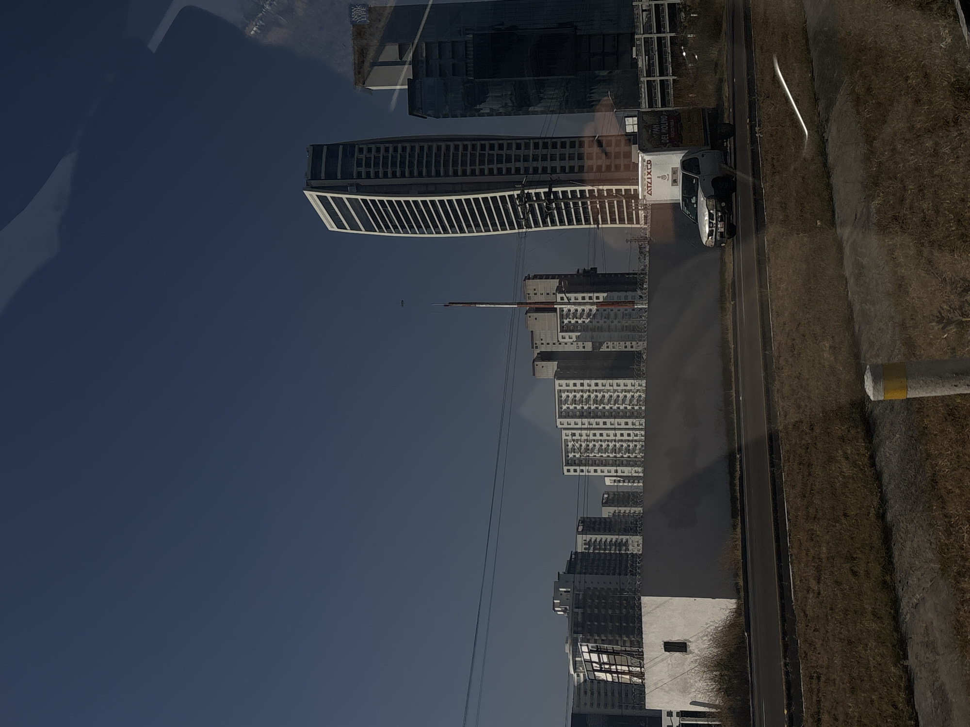The image is a slightly distorted reflection of the city of San Bernardino Tlaxcalancingo, Mexico. Several high-rise buildings of varying designs are prominent, including a curved white building with a grid-like facade. There is also a delivery truck parked near the buildings, labeled with the word "ATZIZXO." The sky is clear and blue. A narrow road runs alongside a strip of overgrown vegetation. The overall view seems to be captured through glass, exhibiting reflections.