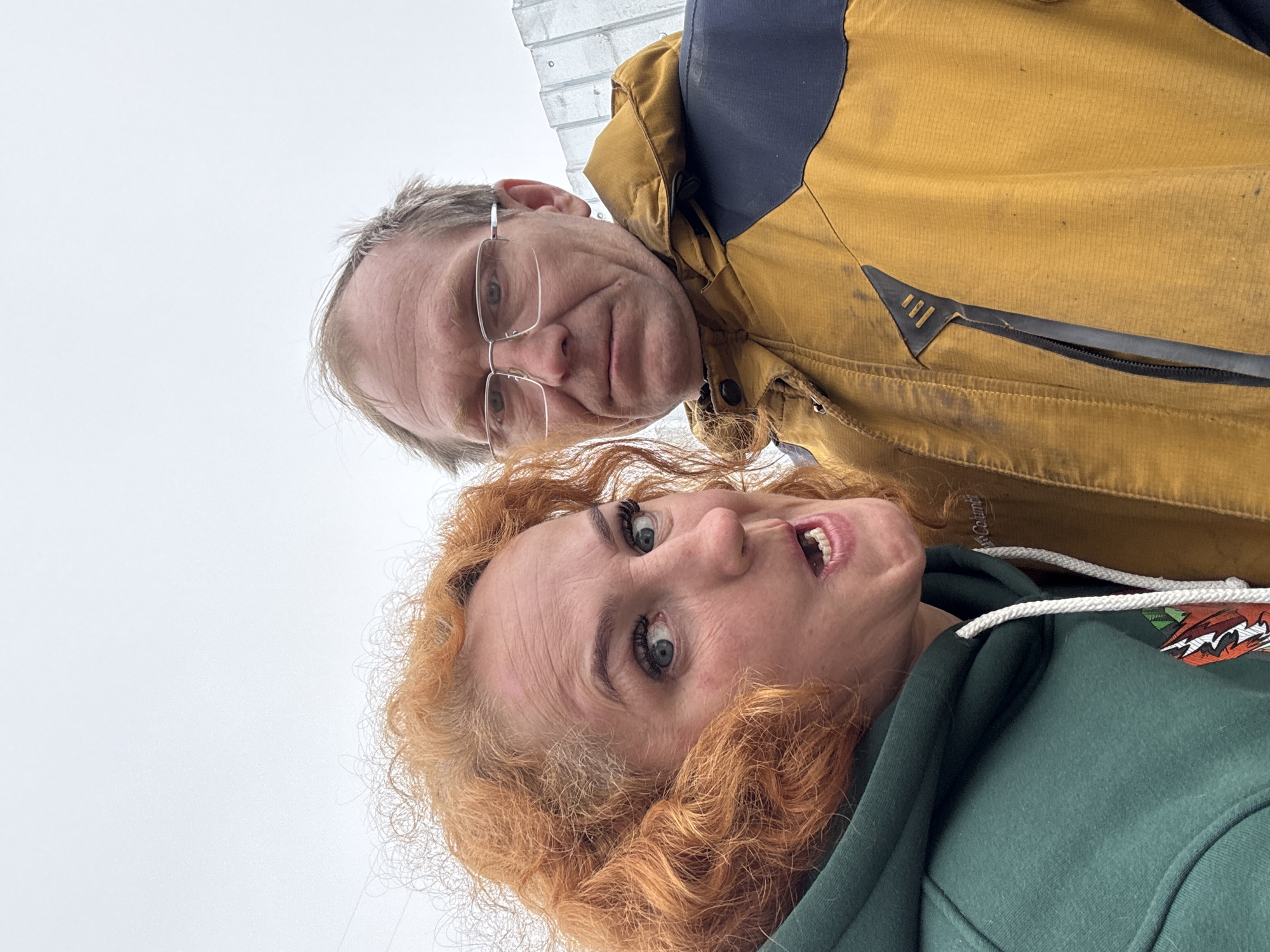 Two individuals, a man and a woman, are positioned in close proximity to each other, facing the camera from an unusual low-angle perspective. The man is older, wearing glasses and a brown jacket with a navy-blue upper section. The woman is younger with red, curly hair and is wearing a green hooded sweatshirt with a drawstring. The background is an overcast sky.
There is no information to confirm the location in the city of Chaikovsky, Russia, or if the photo was even taken there.