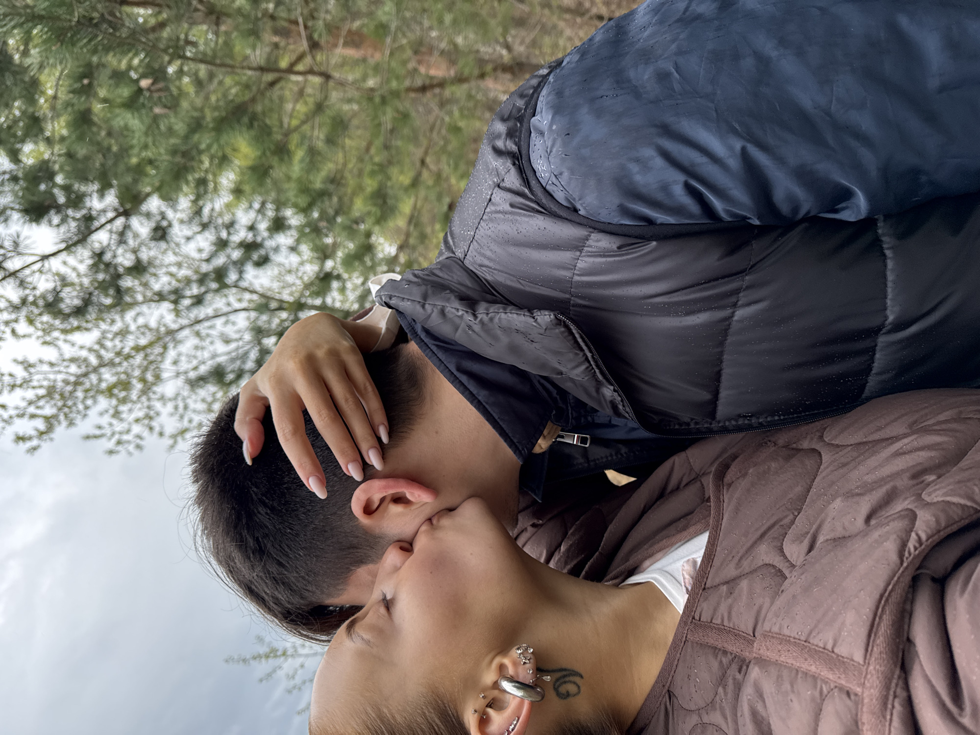 A close-up, slightly angled photograph depicts a couple kissing in an outdoor setting, possibly a wooded area near Chastyye, Russia. The man is wearing a black and blue puffy jacket. His right hand is placed on the back of his head. The woman is wearing a brown puffy vest over a white shirt. She has multiple ear piercings and a tattoo visible near her left ear. The background consists of trees and an overcast sky. Raindrops are visible on their jackets. The image is taken from a slightly lower angle, looking upwards.