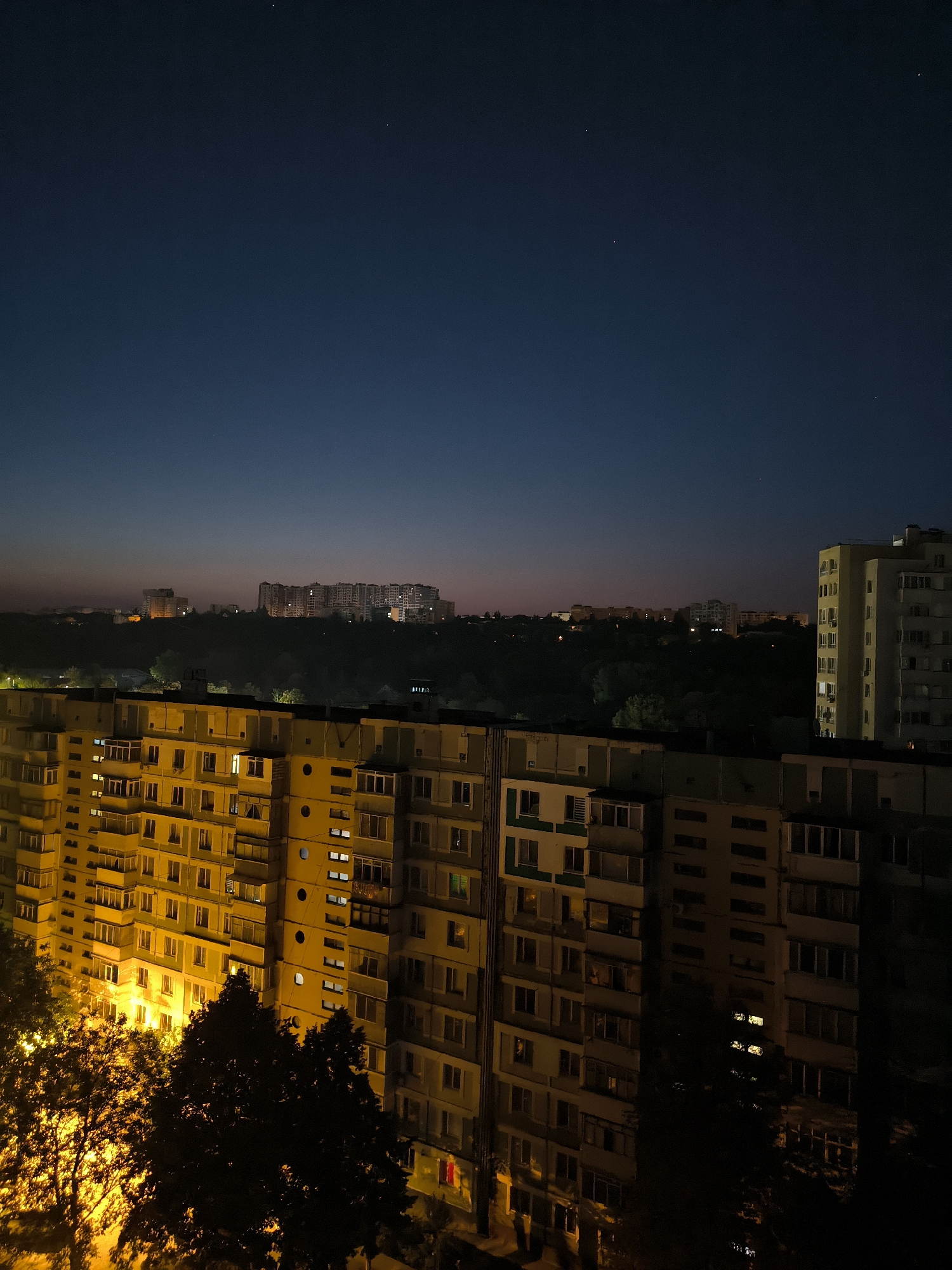The image shows a nighttime view of a residential area in Chișinău, Moldova. The foreground is dominated by a large, multi-story apartment building, illuminated by warm yellow streetlights and interior lights shining from its windows. The building exhibits a common Soviet-era architectural style, characterized by its repetitive balconies and light-colored facade. To the right, another apartment building stands slightly further back, also lit from within. In the background, more apartment blocks and city lights are visible, stretching across the horizon under a dark blue twilight sky dotted with a few faint stars. Dense trees partially obscure some of the lower levels of the foreground building and create dark silhouettes against the illuminated structures. The overall scene depicts a typical urban environment at dusk or early night, with the lights of the city creating a warm glow against the darkening sky.