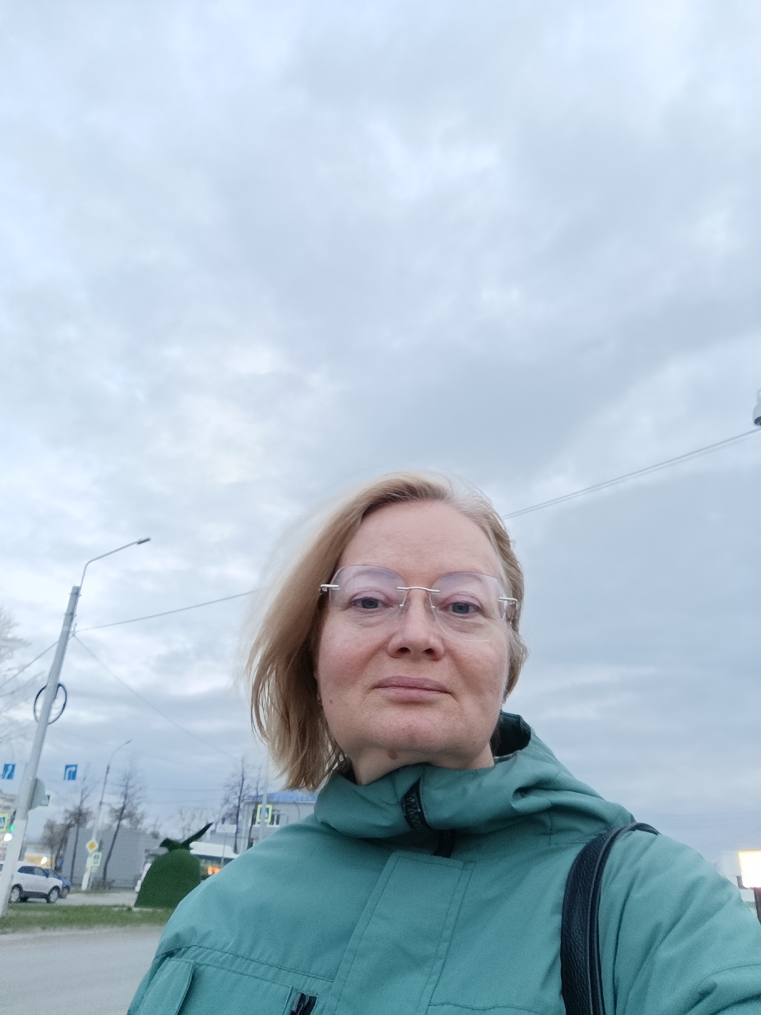 A woman with fair skin and blonde hair is standing outside, presumably in the city of Chaikovsky, Russia. She is wearing glasses and a teal hooded jacket with a black shoulder bag strap visible on her right shoulder. The woman appears to be taking a selfie. Behind her is a street scene with overcast skies. A light pole is to the left, and street signs with blue and white markings are nearby. A white car is parked on the street. A green topiary in the shape of a tank can be seen on the right side.