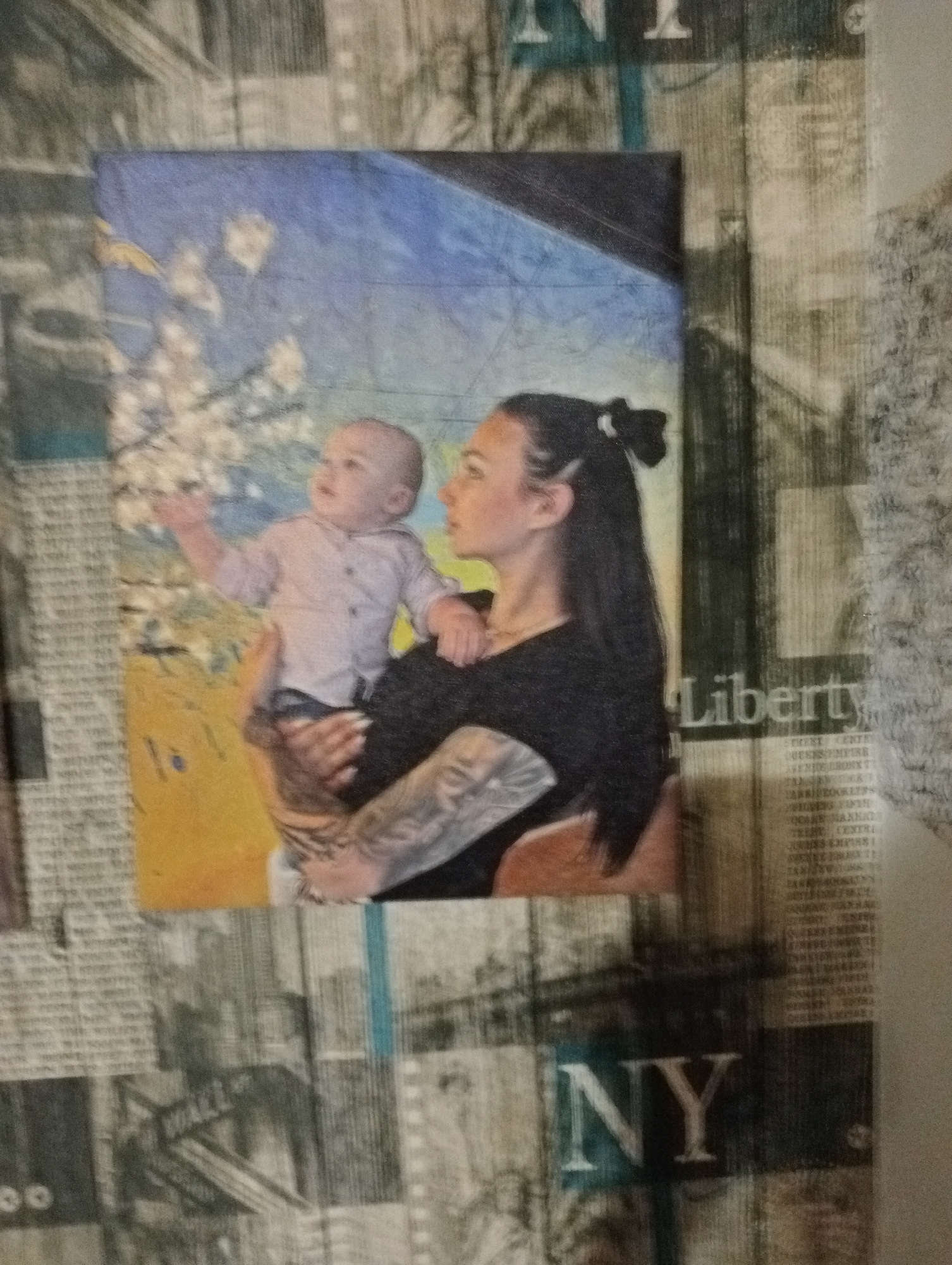 The image shows a painting of a woman holding a baby. The woman has dark hair pulled back in a bun and is wearing a black top. She has tattoos on her arm. The baby is wearing a light-colored shirt and is reaching out its arm. The background of the painting is a mix of blue sky and yellowish-orange ground with white flowers. The painting is framed by wallpaper that features newspaper-style graphics. Visible text includes "Liberty" and "NY" on the wallpaper. The overall impression is a candid or staged portrait of a mother and child. The lighting and colors in the painting suggest it could be daytime, possibly spring due to the flowers. The wallpaper gives a hint of a possible urban setting or New York City theme, but the location of the painting itself is unknown.