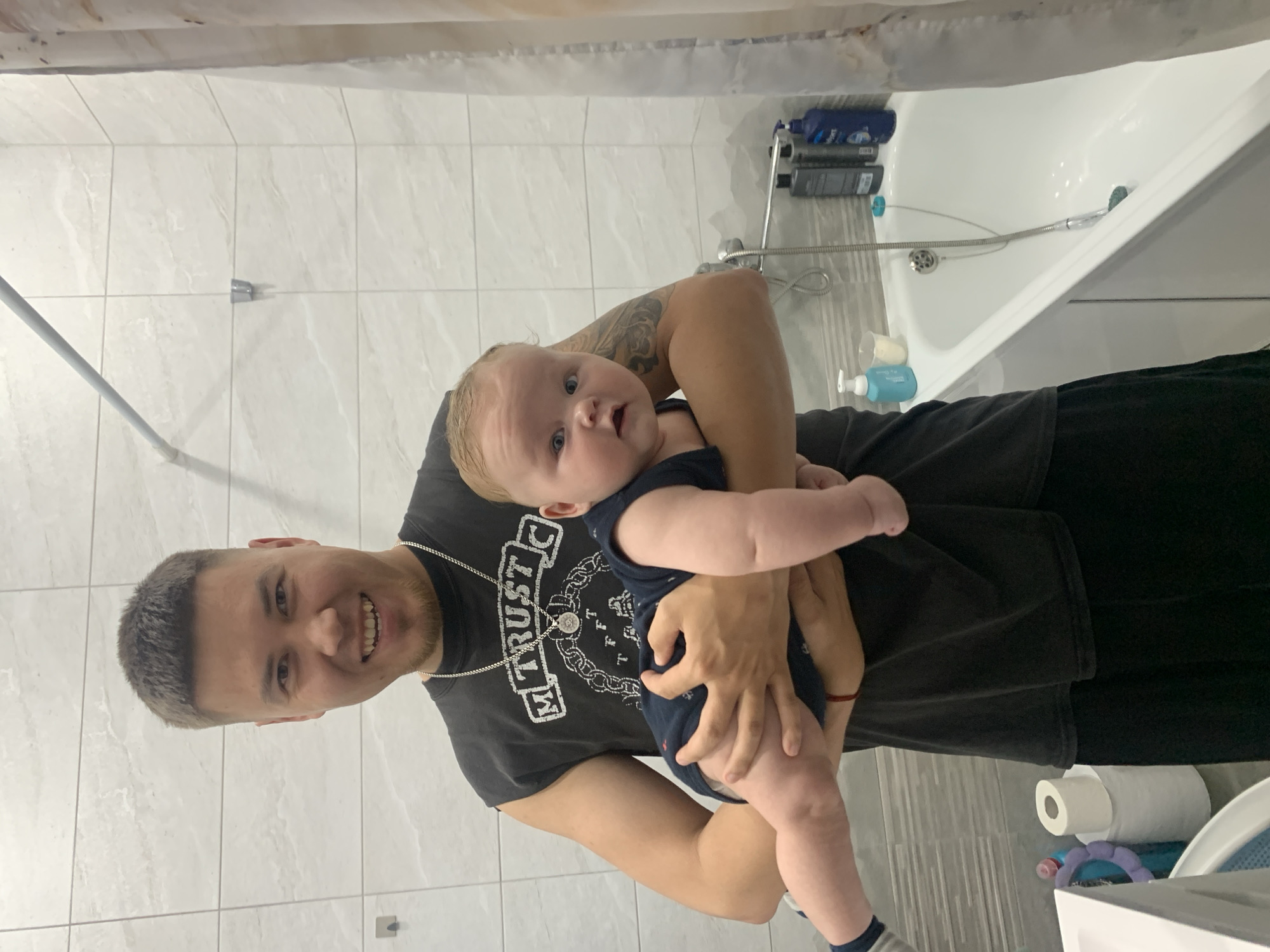A man in a black t-shirt is holding a baby in a bathroom in Stavropol, Russia. The man is smiling and looking at the camera. The baby is looking at the camera with a surprised expression. The bathroom has white tiled walls and a white bathtub. There are several bottles of shampoo and conditioner on the edge of the bathtub. There is also a roll of toilet paper on the counter next to the sink.