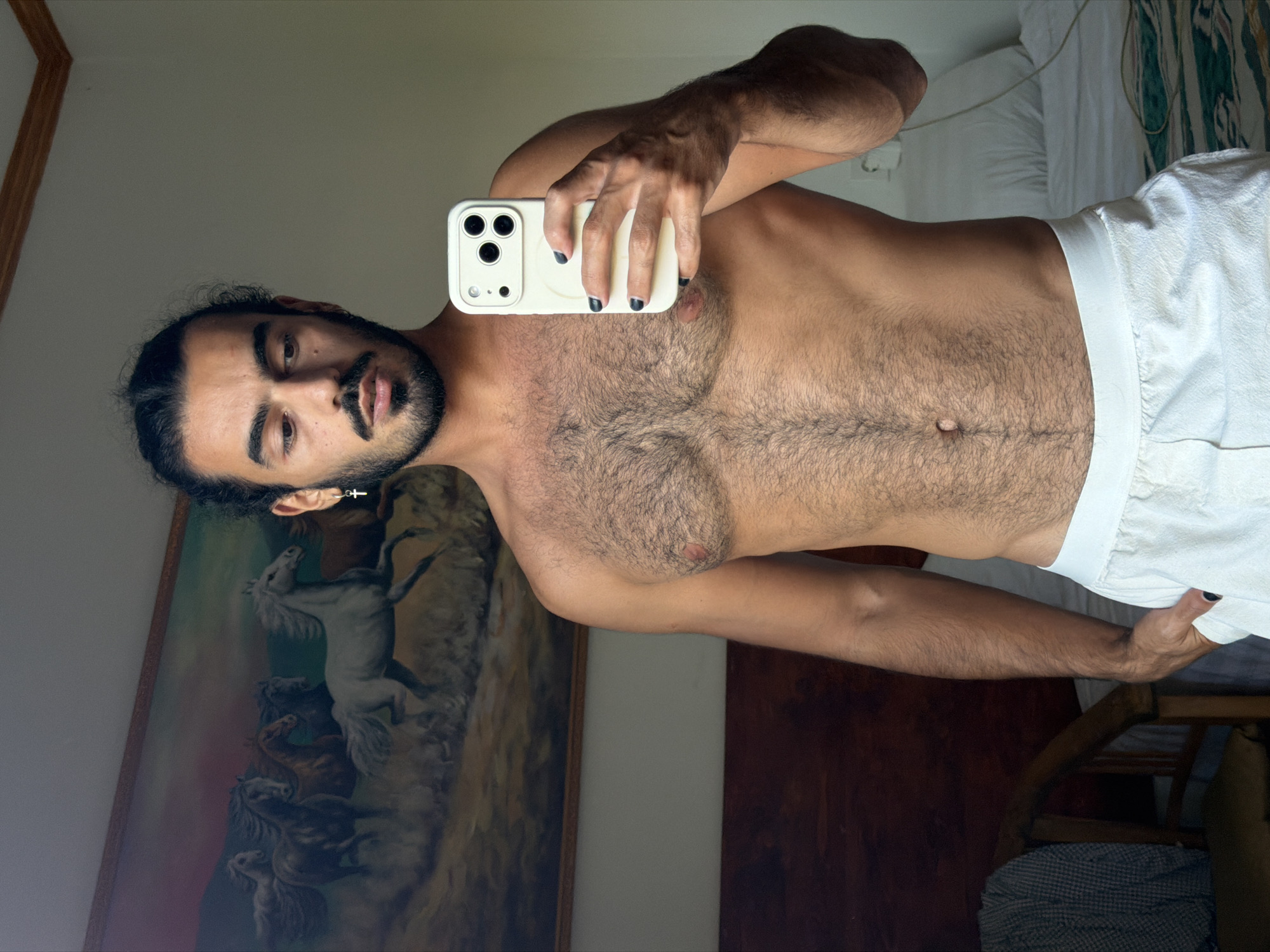 The image shows a man taking a mirror selfie. He is topless, wearing white shorts. He has dark hair, a mustache, and a beard, and is wearing a small silver earring. His fingernails are painted black. In the background, there is a painting of horses and a bed with white sheets. The scene appears to be indoors, possibly a bedroom. The lighting suggests it is daytime.