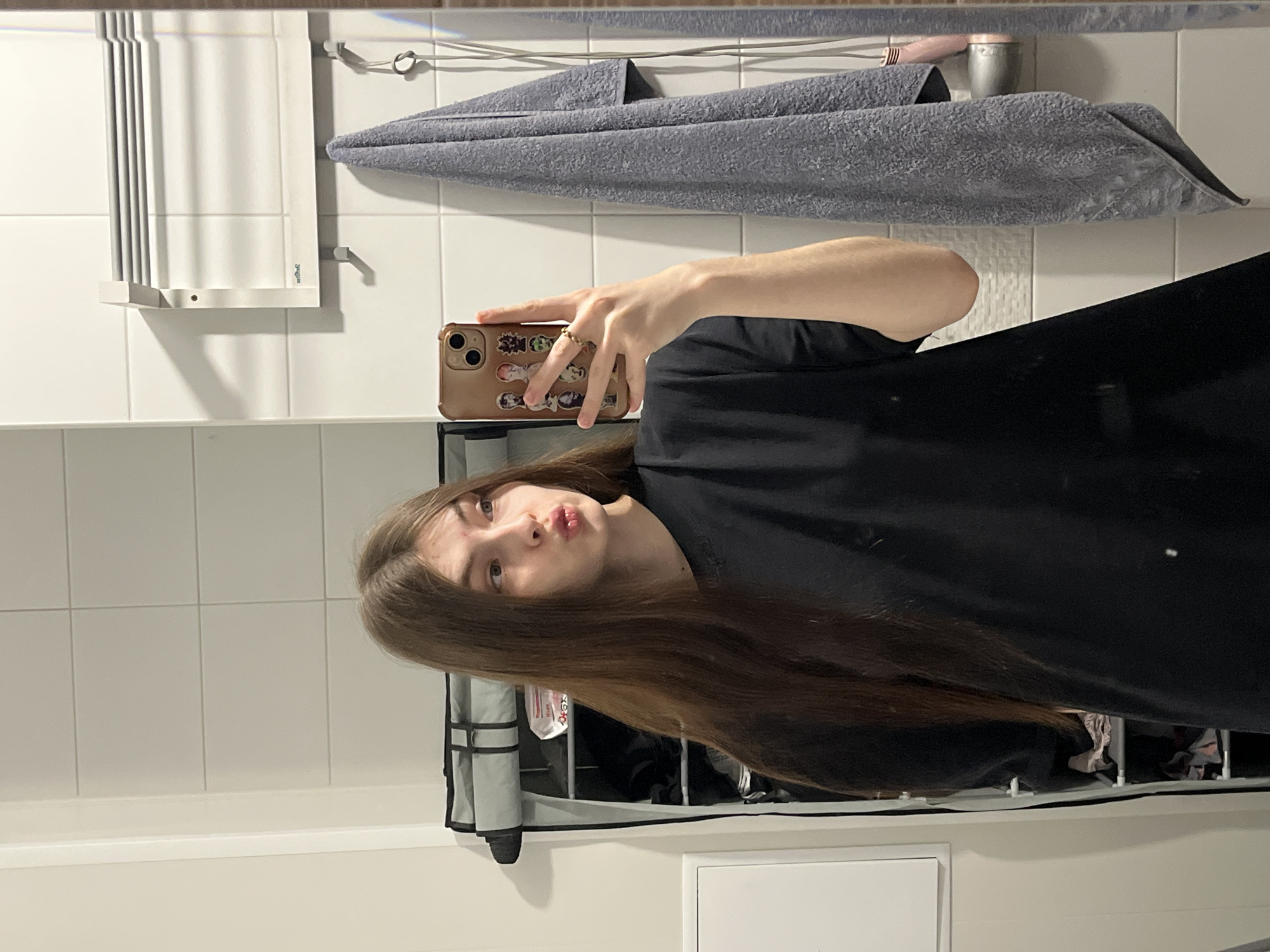 A person is taking a selfie in a bathroom. The bathroom has white tiles, a gray towel hanging on a rack, a mirror, and a storage unit. The person has long, brown hair and is wearing a black shirt. The person is holding a phone with a brown case. The phone case has stickers on it. The location is unknown, but the query specified Saint Petersburg, Russia.