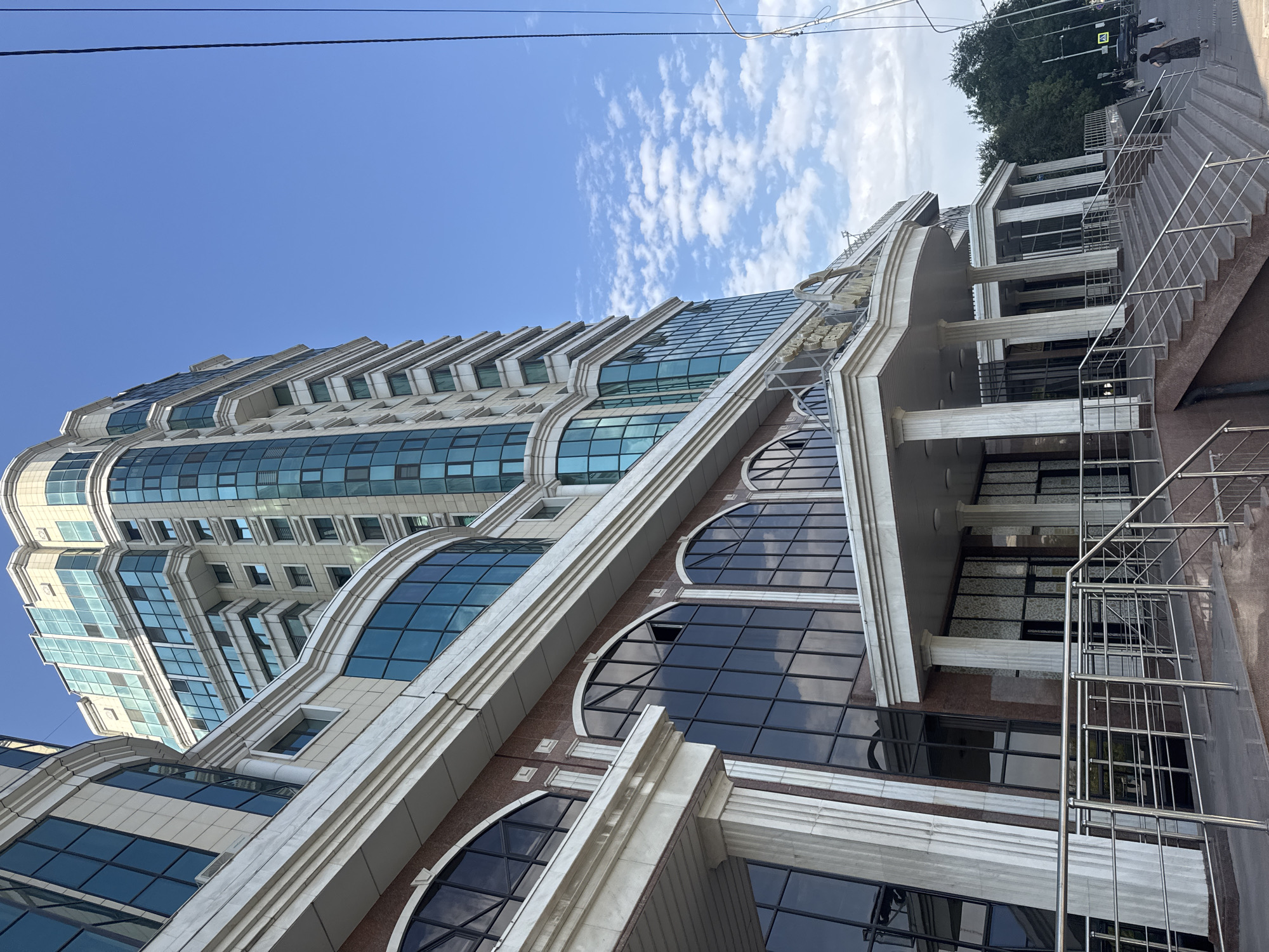 A wide-angle, low-angle, exterior shot captures the modern facade of a multi-story building in Almaty, Kazakhstan, on a clear day with scattered clouds. The building features a mix of light beige stone and dark, reflective glass, with rounded architectural elements on the lower floors and a more angular design on the upper floors. The entrance area showcases classical-style columns supporting a canopy, with a wide set of stairs leading up to it. In the foreground, a metal railing lines the stairs and a walkway. To the right of the building, a few trees are visible, along with a street sign and a black car. Several pedestrians are walking on the sidewalk in the distance. The sky is a vibrant blue, with fluffy white clouds, and power lines are visible on the right side of the frame. The building's architecture suggests a commercial or residential complex.