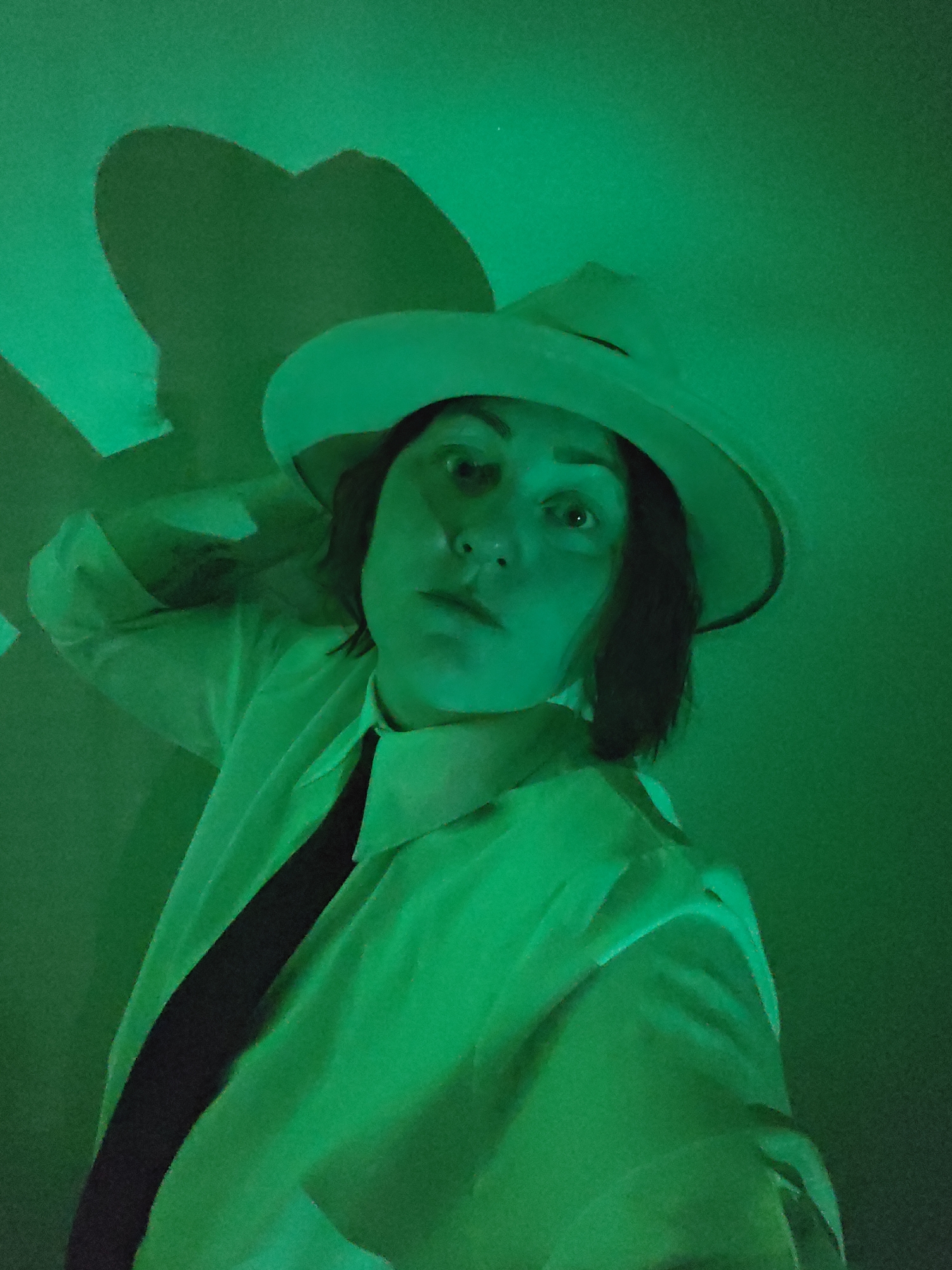 A person wearing a white shirt, black tie, and white hat is illuminated by a green light source. The person's hand is raised near their head. A shadow of the hat is visible on the wall behind the person. The scene is indoors. There is no information about the location of this image in Markovskii, Russia.