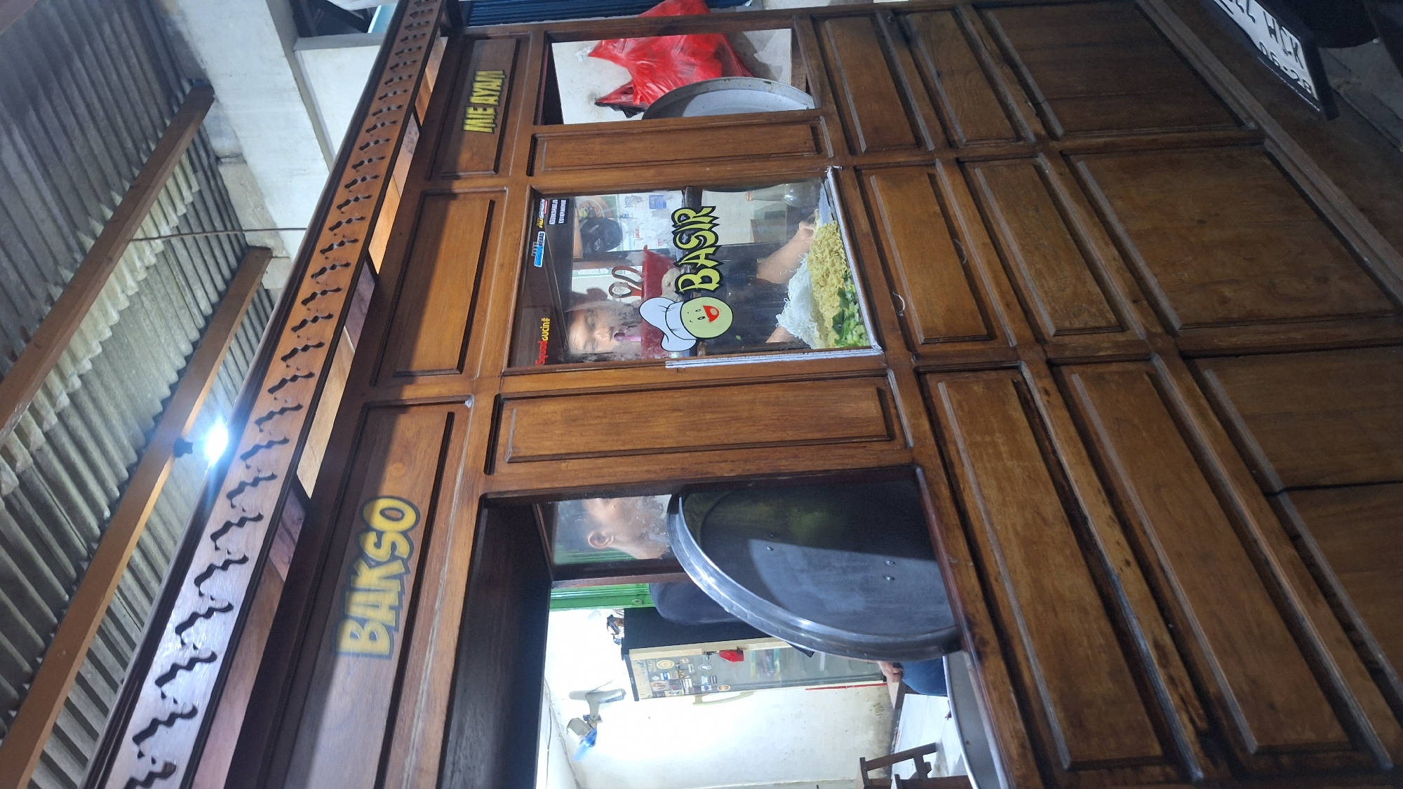 The image shows a food stall in Sukoharjo, Indonesia. The stall is made of dark wood with several serving windows. The window on the left has the word "BAKSO" written vertically. The middle window has a sticker with "BASIR" and a cartoon chef. Through the windows, a person is visible serving food, likely noodles. The background suggests an open-air structure with a corrugated metal roof. A license plate is seen affixed at the top right.
