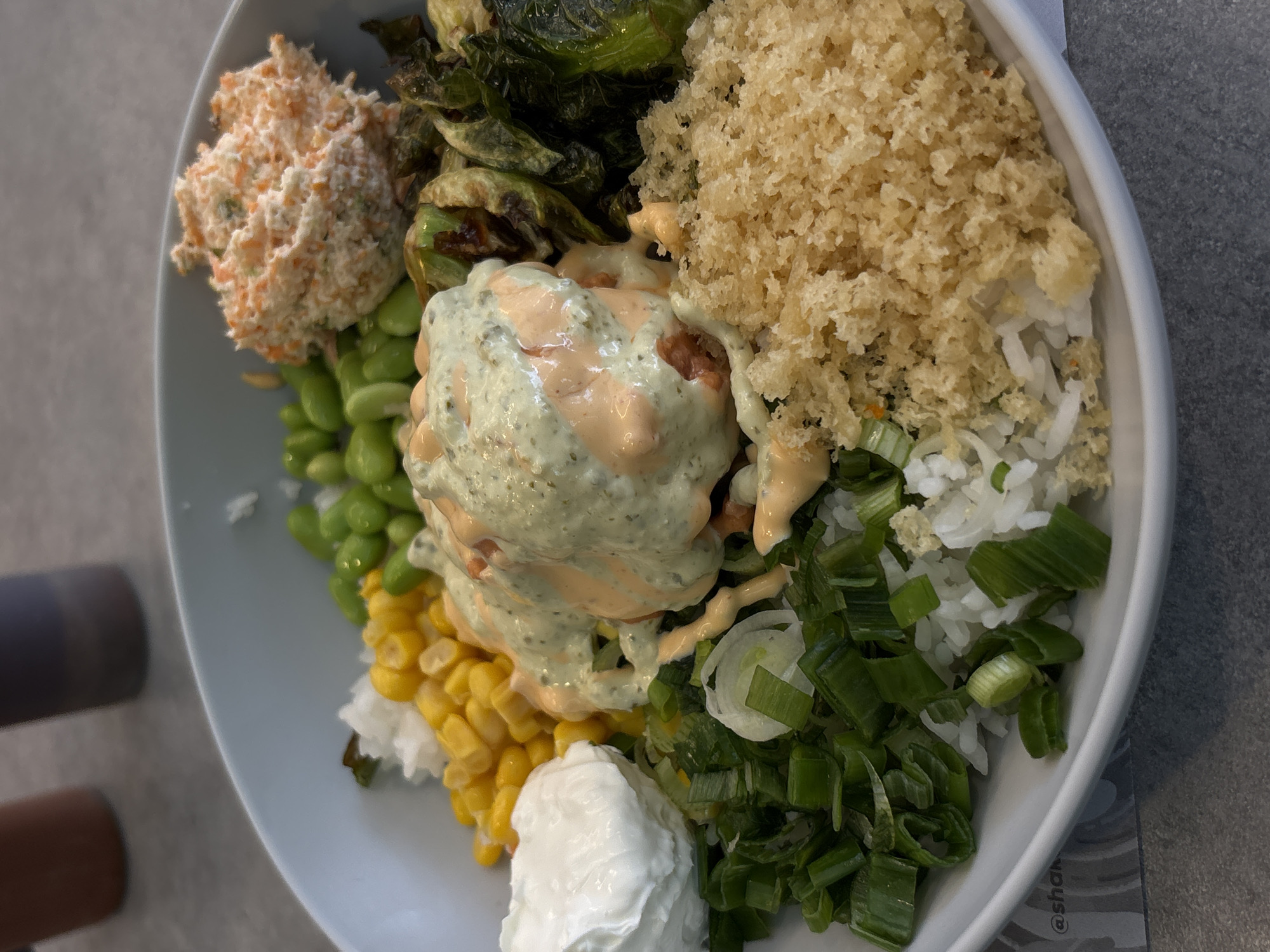 This close-up, overhead photograph showcases a colorful and varied food bowl, likely a custom rice bowl or poke bowl, presented in a casual dining setting in Mexico City, Mexico. The light-colored bowl rests on a grey, textured surface.
The bowl is packed with diverse ingredients. In the top left, there's a scoop of orange-hued, creamy shredded salad, possibly a carrot salad or slaw. Adjacent to it are several dark green, roasted or sautéed Brussels sprouts.
At the center of the bowl, a main protein, possibly pieces of fish or chicken, is lavishly drizzled with two sauces: one creamy, light green sauce (perhaps herb or avocado-based) and another orange or pinkish sauce, which might be spicy. To the top right, a large pile of light brown, crispy tempura flakes or panko breadcrumbs adds texture.
Beneath these toppings, white rice forms the base, particularly visible on the right side, topped with a generous scattering of freshly chopped green onions. Towards the bottom center, a dollop of white creamy substance, likely sour cream or a similar dressing, sits next to a portion of bright yellow corn kernels. Green edamame beans are also visible on the left side of the bowl.
The food appears fresh and well-composed. Faint text, "@shan", is visible on the surface beneath the bowl in the bottom right corner, suggesting a brand name or social media tag. No individuals are present, and the time of day or weather conditions are not discernible from the image.