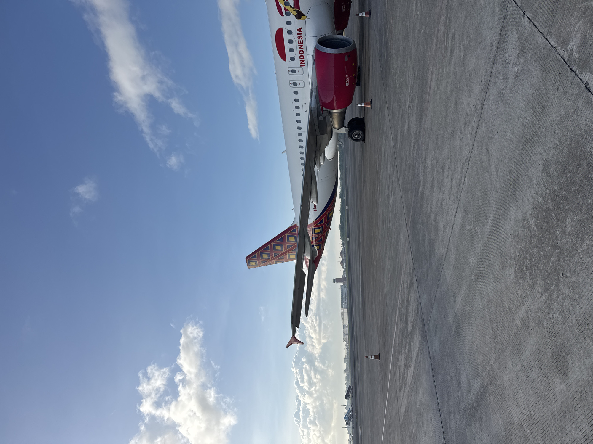A daytime shot depicts an airplane on a tarmac with a blue sky overhead interspersed with white clouds. The airplane is painted white with red accents and the word "INDONESIA" is visible on its side. The tail fin features a colorful, patterned design. The airplane is positioned on a concrete tarmac. Orange cones are placed along the tarmac. A control tower and other buildings are visible in the distance. The location is Unknown, Unknown.