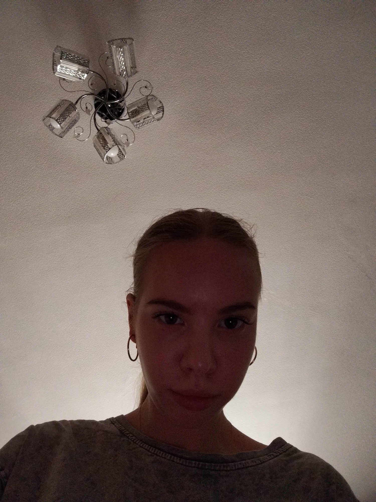 This is a medium shot selfie of a young woman with blonde hair pulled back, looking directly at the camera. She is wearing a grey t-shirt and large hoop earrings. Her expression is neutral. The background is a textured white ceiling with a decorative light fixture. The lighting is dim, with a warm tone, suggesting it may be evening or indoors. The image is framed from the chest up, with the ceiling fixture visible in the upper portion of the frame.