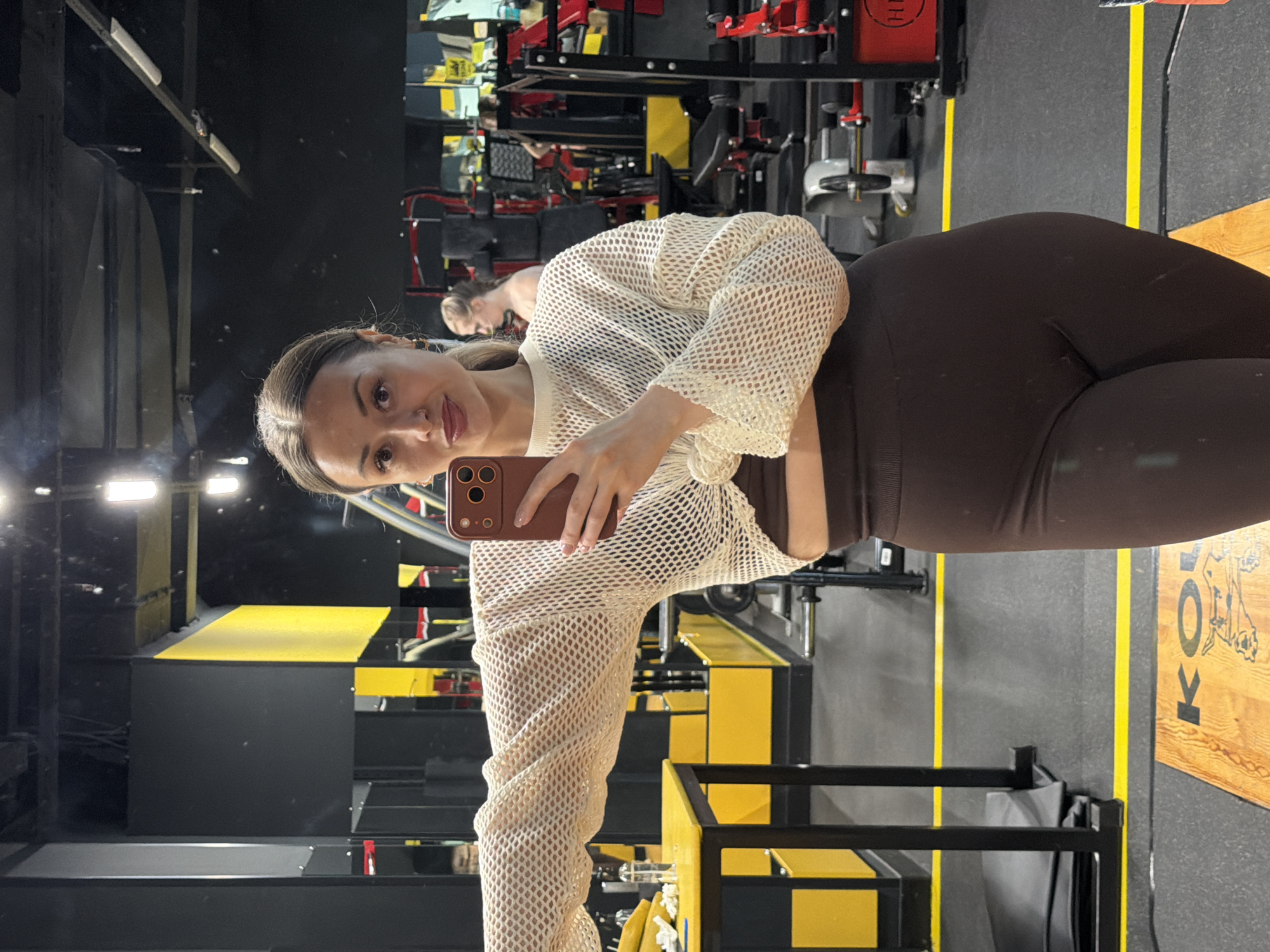 A woman is taking a selfie in a gym. She is wearing brown leggings and a beige netted long-sleeved top, and holding her phone in her right hand. Other gym equipment and another person can be seen in the background, reflected in a mirror. The scene appears to be indoors. I am unable to determine if this is in the city of Krasnoyarsk, Russia.