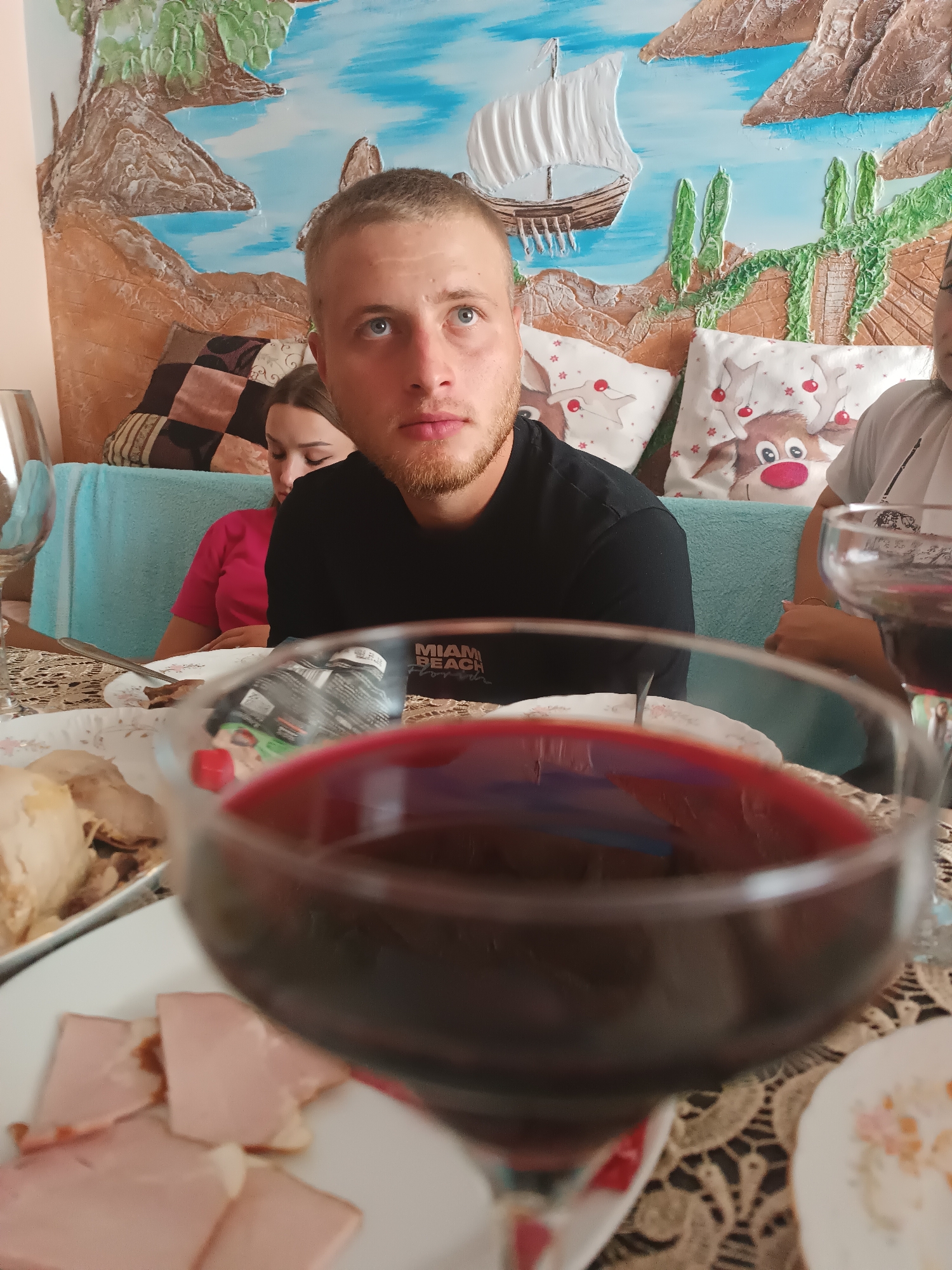 The image depicts an indoor scene with a family gathering, possibly in Durlești, Moldova. In the foreground, a wine glass filled with red wine is prominently displayed, its contents blurring the background. Behind the glass, on a table laid with a lace tablecloth, are plates of food, including sliced ham and what appears to be roasted meat. The background features a man with light blue eyes and a beard, looking upwards, wearing a black t-shirt with "MIAMI BEACH" printed on it. To his left, a young woman in a pink shirt is looking down. Behind them, a light blue couch is visible, adorned with cushions featuring cartoon reindeer. The wall behind the couch is decorated with a textured mural depicting a seascape with a sailing ship, rocks, and greenery. Another person is partially visible on the far right, also holding a wine glass. The overall atmosphere suggests a casual meal or celebration.