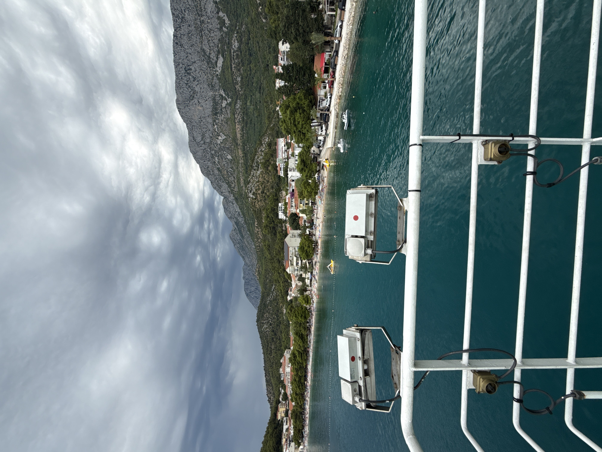 A photograph taken from the deck of a vessel, looking towards the coast of Drvenik, Croatia. The water in the foreground is a vibrant turquoise, with white railings and two large, metal floodlights visible. The floodlights have black cables attached and appear to be mounted on a metal frame. Beyond the railing and lights, a beach is visible, lined with trees and buildings. The beach is populated with numerous people, some of whom are sunbathing or swimming. Several small boats are anchored in the water. The town is nestled at the base of a steep, rocky mountain covered in dense green vegetation. The sky above is overcast with dramatic, dark clouds. The overall impression is of a coastal town on a cloudy day, viewed from the sea.