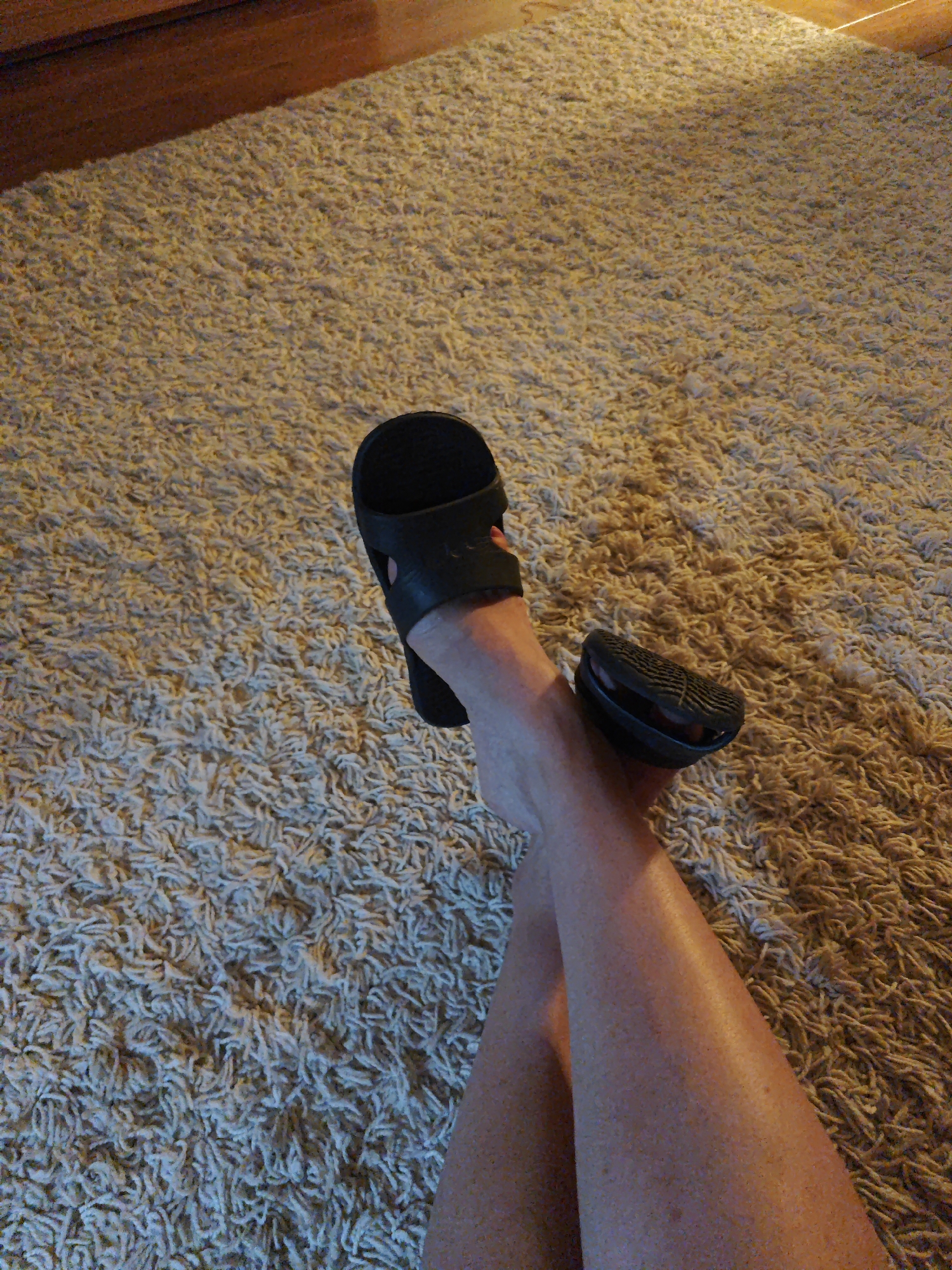 The image shows a pair of legs and feet resting on a shag carpet. The legs are bare and appear to be lightly tanned with freckles. One foot is wearing a black slide sandal, and the other foot is also wearing a black slide sandal, but it is positioned slightly off the foot, revealing the sole. The carpet is a light tan or beige color and has a thick, fluffy texture. In the upper left corner of the image, a wooden surface is visible, possibly a piece of furniture or trim. The lighting in the image suggests it might be indoors, possibly in the evening, given the warm glow cast on the carpet. There is no visible text in the image. The overall mood is relaxed and casual.