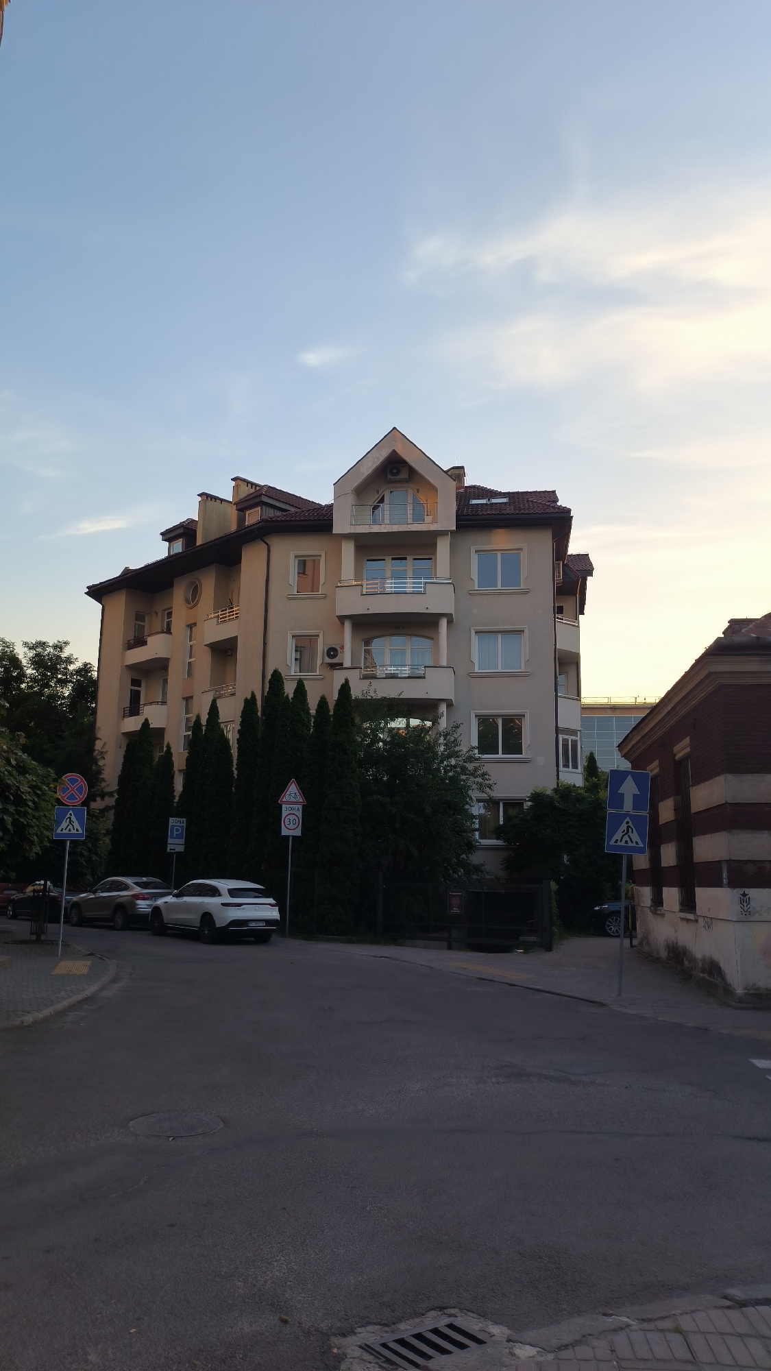 This is a photograph of an apartment building and street scene in Lviv, Ukraine. The building is modern, with a light-colored facade, multiple balconies, and a red-tiled roof. Several cars are parked along the street in front of the building, including a white SUV and a silver sedan. There are several traffic signs visible, including a pedestrian crossing sign and a speed limit sign. Trees and bushes line the street, and the sky is a pale blue with some wispy clouds, suggesting it is either early morning or late afternoon. The overall impression is one of a typical urban residential street.