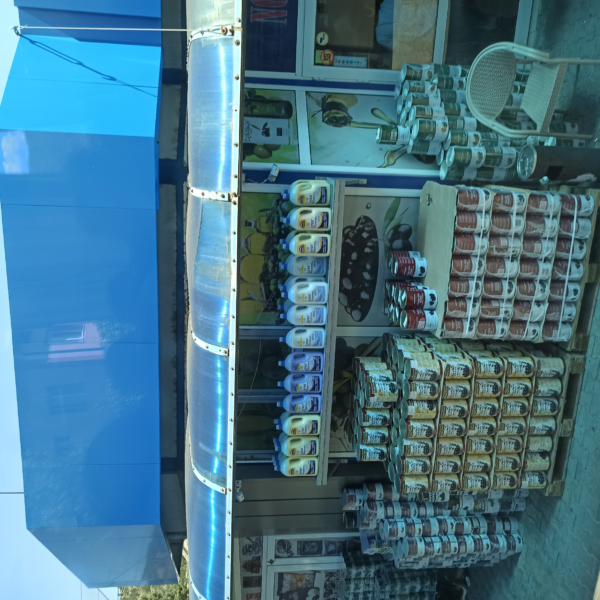 This image depicts an outdoor market stall in Kresna, Bulgaria. The stall has a blue, box-like structure covering the top and a transparent, ribbed awning extending outwards. Displayed on shelves are numerous bottles of cleaning detergent, with some labeled "lenor" in yellow and purple. Below the shelves, a large quantity of canned goods is stacked on pallets. These cans appear to contain olives, with labels featuring green olives and text indicating "black olives" and "green olives." A variety of other canned goods are also visible, some in white and green packaging and others in dark red and white. To the left, more canned goods are stacked high. A white plastic chair and a metal trash bin are also present. The background shows a building facade with blue siding. The scene is brightly lit by natural daylight.