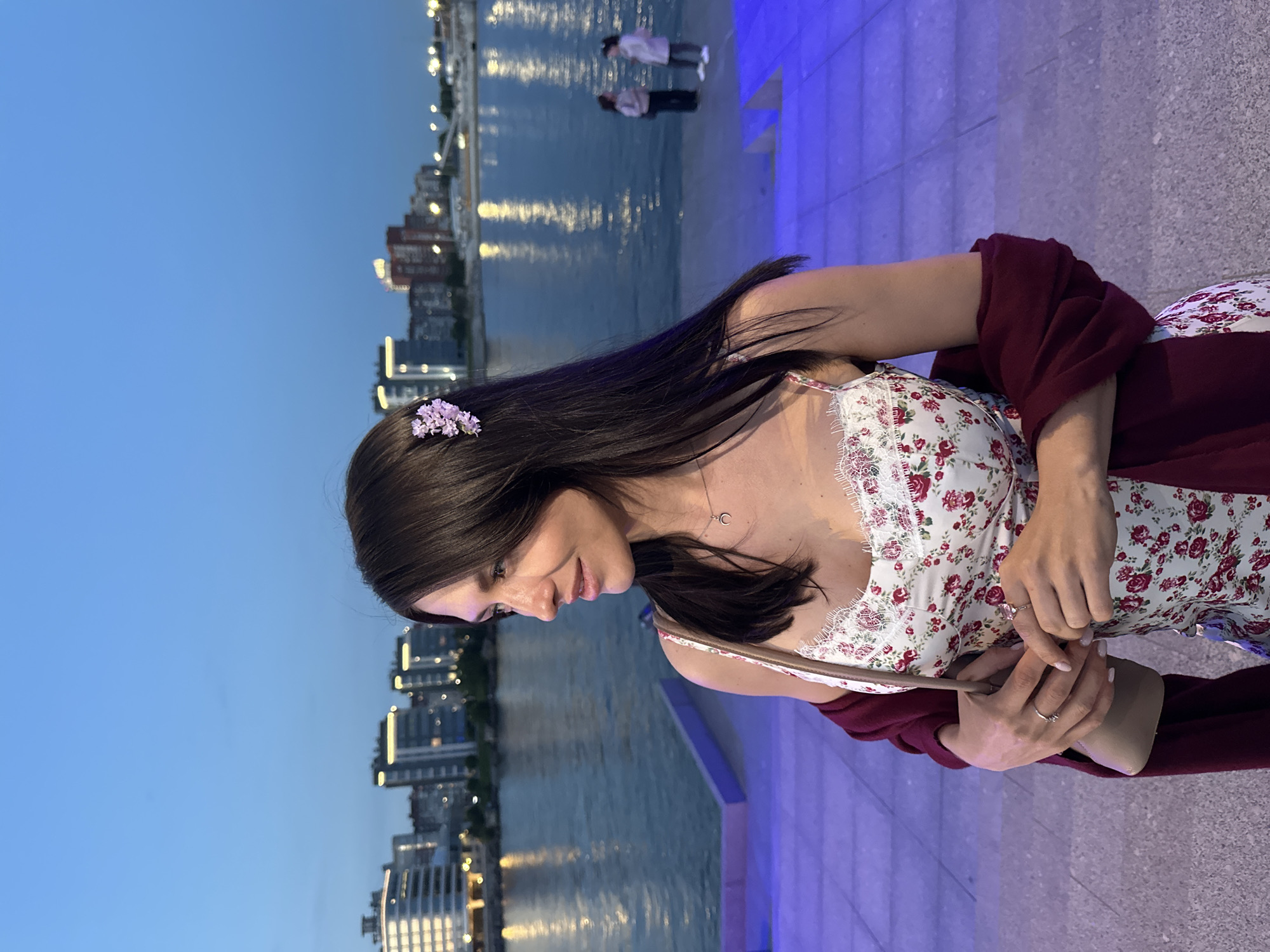 The image depicts a young woman standing on illuminated blue steps near a waterfront in Saint Petersburg, Russia, at dusk. She has dark hair adorned with a small cluster of purple flowers and is wearing a floral print dress with lace trim and a dark red shawl draped over her shoulders. She is holding a small tan handbag and looking off to her right. In the background, the Neva River is visible, with several buildings and lights reflecting on its surface. Two other individuals, a woman and a girl, are standing further away on the waterfront.