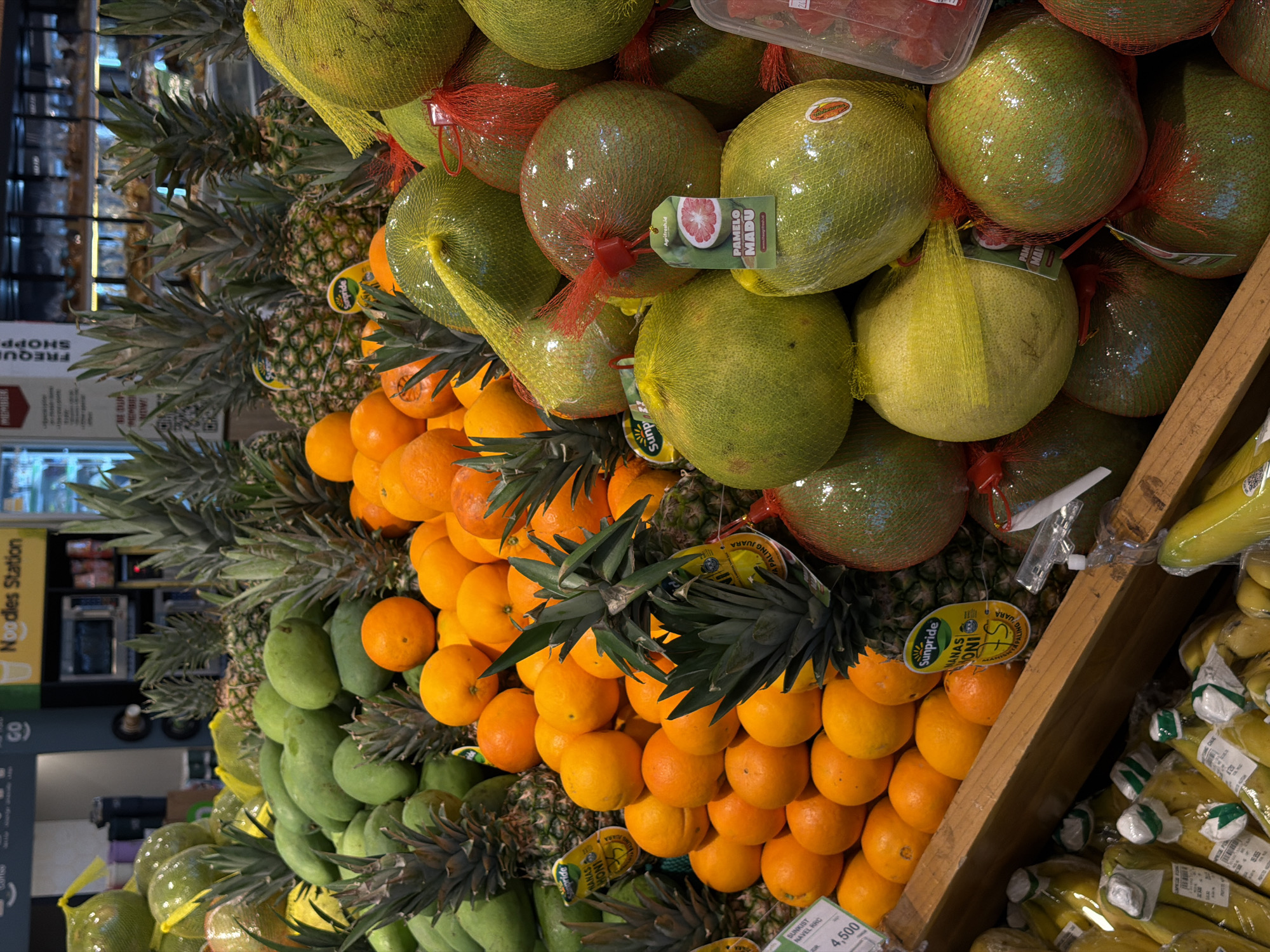 The image shows a display of various fruits at a grocery store, likely in Kuta Selatan, Indonesia. Stacked from left to right are green mangoes, pineapples, oranges and pomelos encased in netting. Bananas wrapped in plastic are visible on the far right, and a wooden support structure holds the arrangement. Price tags are visible among the fruit, and store signage is visible in the background, possibly indicating a "Noodles Station".