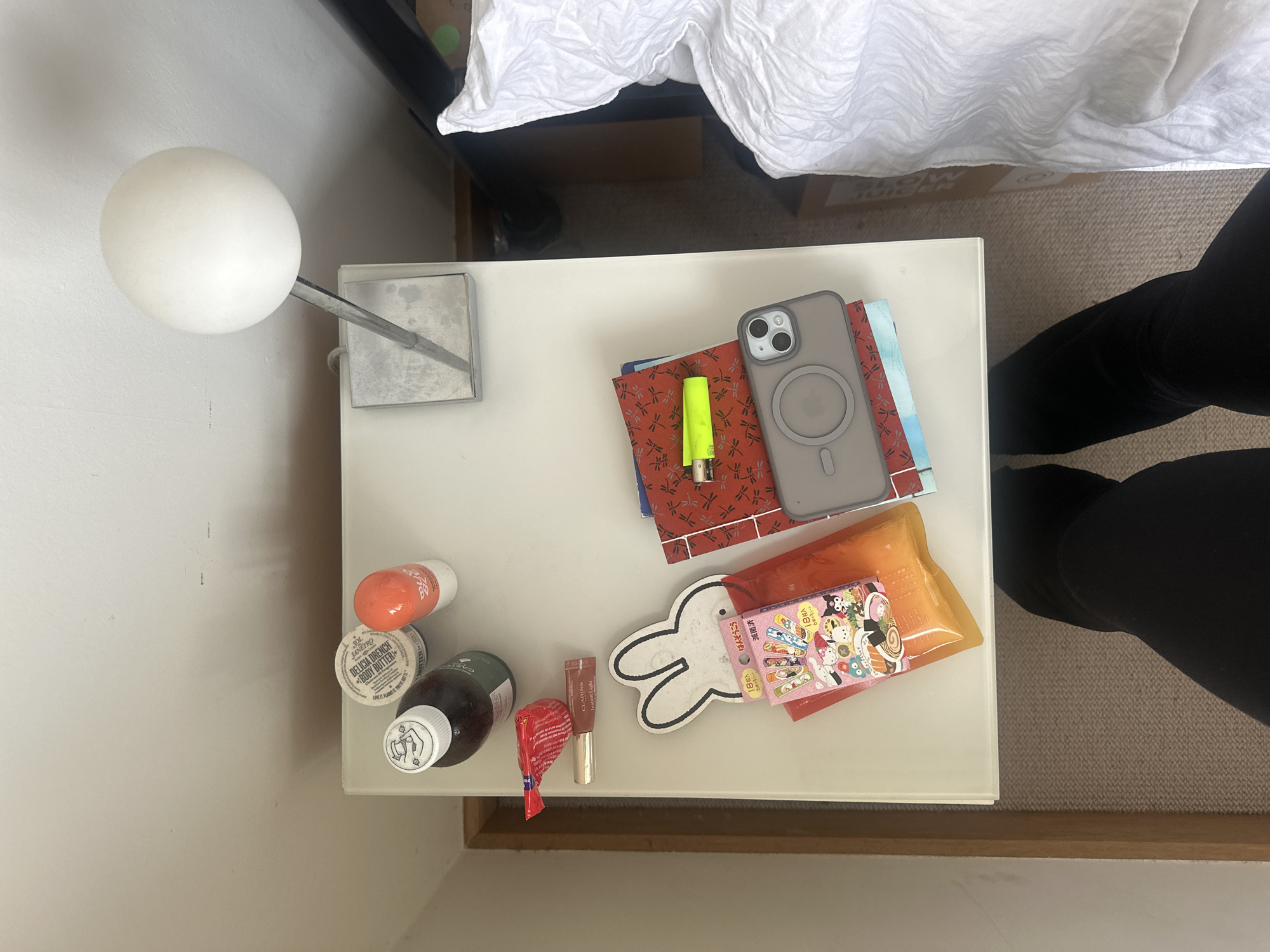 The scene depicts a white bedside table in a room in the city of Wealden, United Kingdom. On the table, from left to right, are a dark brown bottle with a white screw cap, a white cylindrical container of "SOL DE JANEIRO DELICIA DRENCH BODY BUTTER," and an orange cylindrical dispenser. Below these, a small red wrapper lies next to a tube of "CLARINS Instant Light" lip product. A white Miffy-shaped cutout is positioned over an orange flexible packet, which is partially obscured by a small rectangular package labeled "18枚入" (18 pieces) and "はみがくろ" (Hamigakuro) featuring various Sanrio characters as sushi. To the right of these items, a white spherical lamp head is attached to a tarnished silver-toned metallic base. Below the lamp, a stack of items includes a red notebook with a dragonfly pattern, upon which a neon yellow disposable lighter rests. A grey smartphone, encased in a grey MagSafe case, is placed on top of the notebooks. The table rests on a light-colored, flecked carpet. A light beige wall with a wooden trim is visible to the left. To the right, a bed with white, textured bedding and a black bed frame is partially visible. The lower portion of the image shows a person's legs, clad in black trousers or leggings, positioned in front of the bedside table. No discernible actions or interactions are occurring.