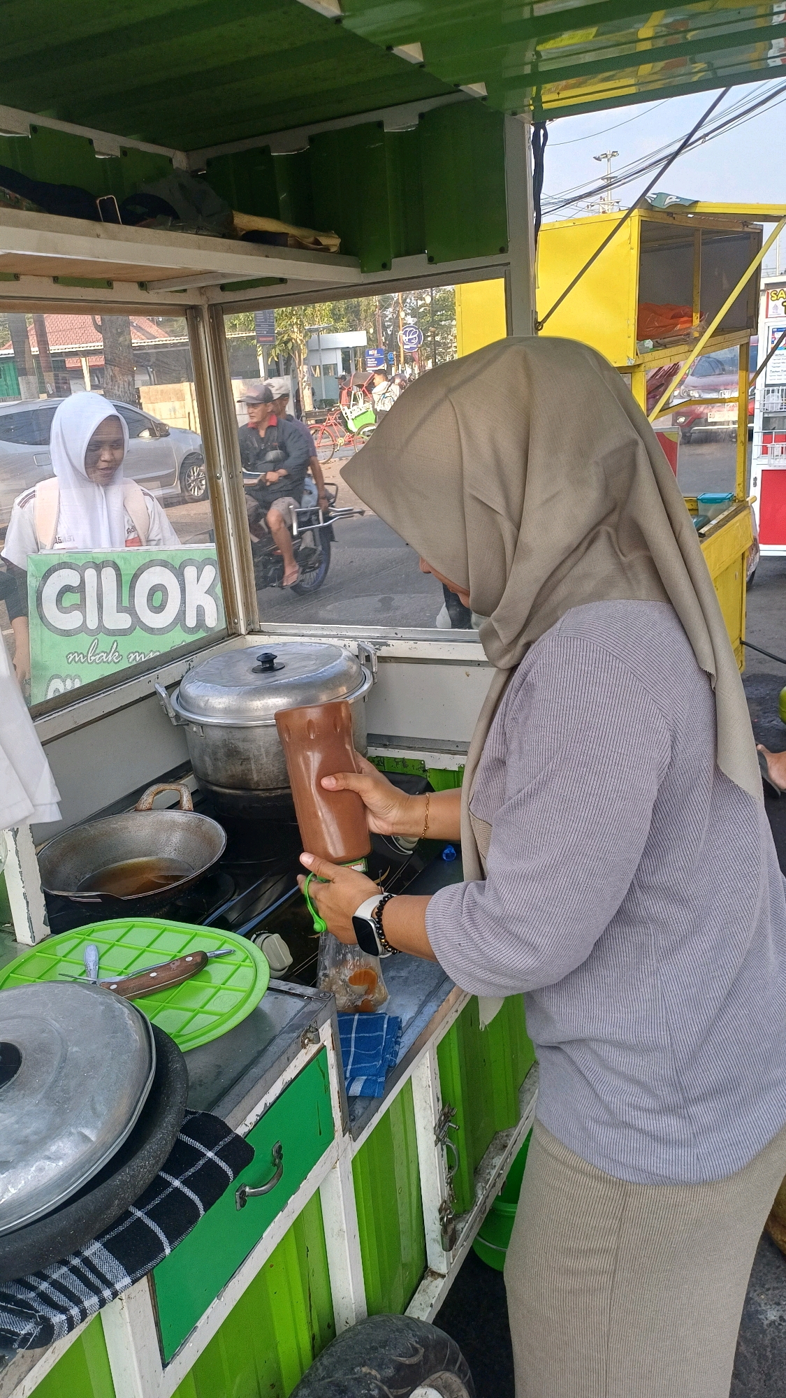 This is a medium shot of a street food vendor preparing food in Klaten, Indonesia. The vendor, wearing a light brown hijab and a grey ribbed shirt, is holding a brown squeeze bottle with a green cap, squeezing sauce into a plastic bag. A young woman wearing a white hijab and a white and grey striped shirt is visible in the background, looking at the vendor. She appears to be a customer. The food cart has a green and white exterior. Various cooking utensils and food items are visible on the cart, including a large metal pot, a frying pan with oil, and a green perforated mat with a knife. In the background, there is street traffic, including a motorcycle, and a yellow food cart. The scene appears to be outdoors on a street, likely during daytime.