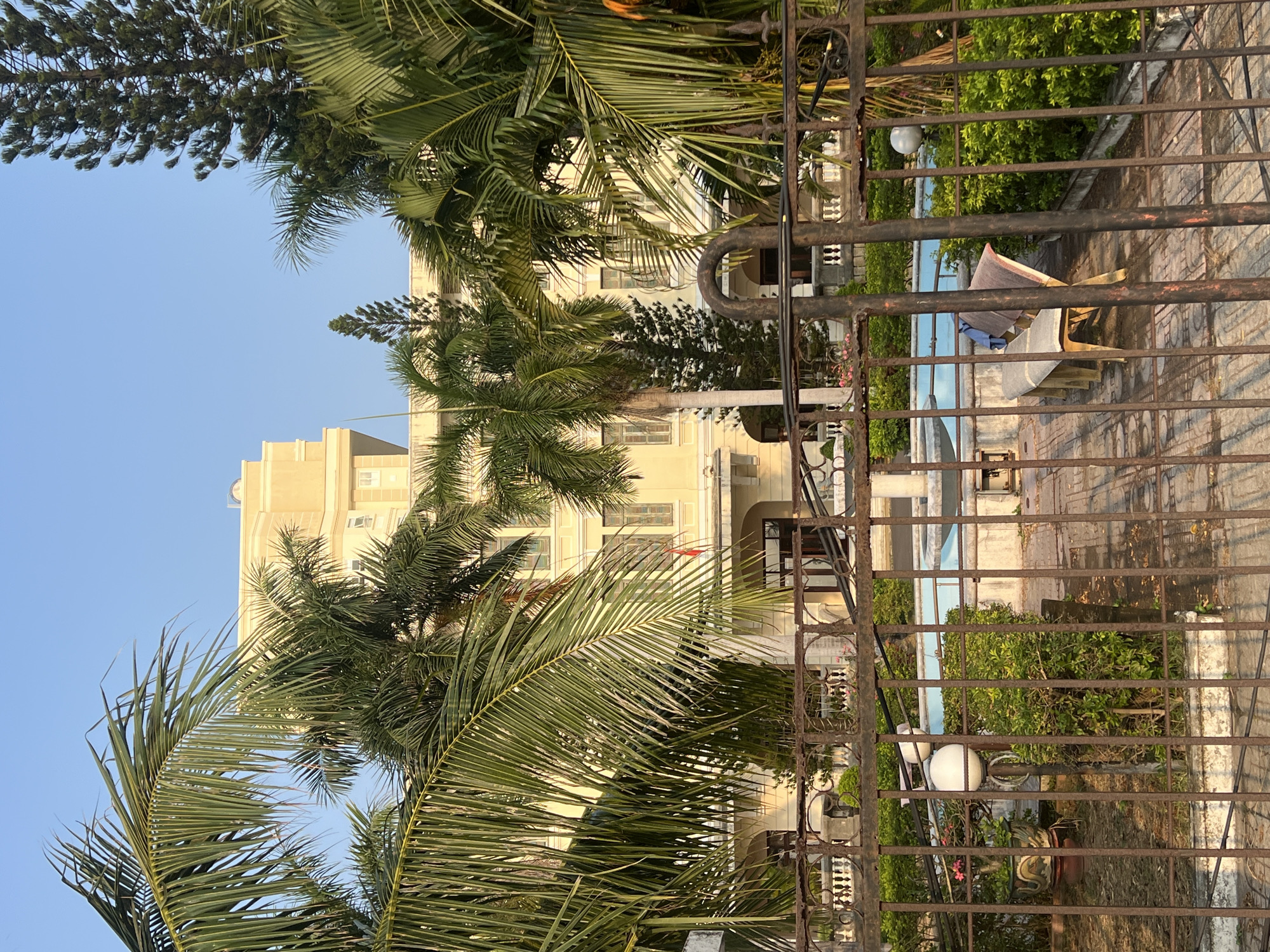 The image, taken in Nha Trang, Vietnam, shows a gated view of a multi-story, light yellow building. The foreground is dominated by a dark, rusty metal gate with vertical bars. Behind the gate, there is a patio or outdoor area with plants, a white chair with some fabric draped on it, and a pool. Palm trees with long, green fronds are visible, partially obscuring the building. The sky is a clear, light blue. The building has multiple windows and some architectural details, including a stepped upper section. There is also some shrubbery visible between the gate and the patio area.