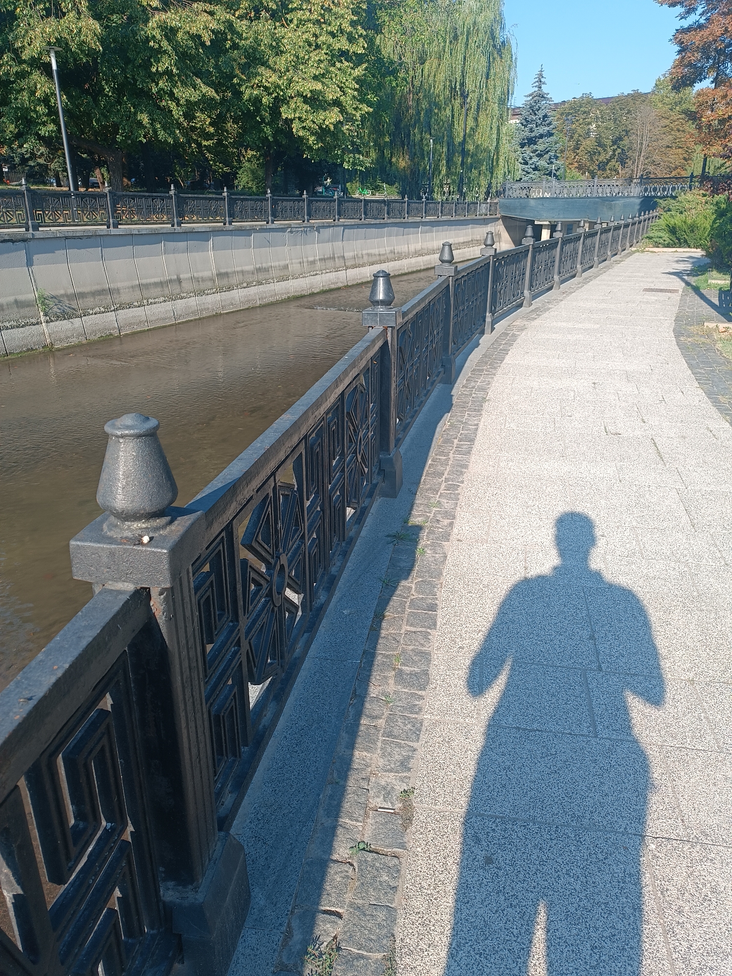 The image depicts a sunlit scene along a paved walkway next to a canal. A dark metal fence lines the walkway, separating it from the canal. The canal water is murky brown, and its banks are lined with concrete. Trees, with green foliage, line the opposite side of the canal. In the distance, a bridge with a decorative railing crosses the canal. The walkway is made of light-colored paving stones, and a distinct shadow of a person is cast across it, indicating the sun is to the left and behind the viewer. The overall environment suggests a park or a promenade area within a populated setting.