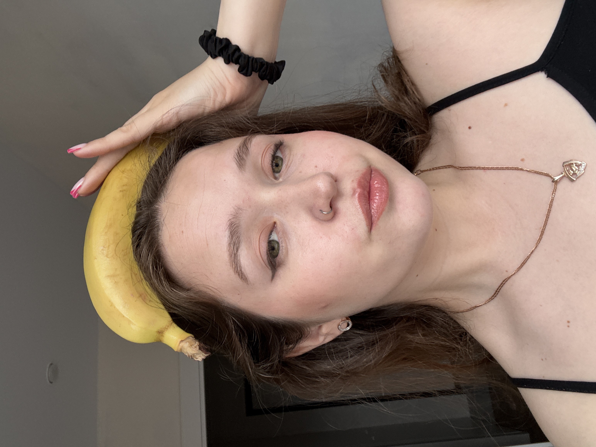 A young woman is holding a banana above her head. She is wearing a black spaghetti-strap top and a gold necklace. She has a nose ring and is wearing pink nail polish. Her hair is brown and wavy. The background appears to be a plain wall. There is no clear indication of the location being Izhevsk, Russia.