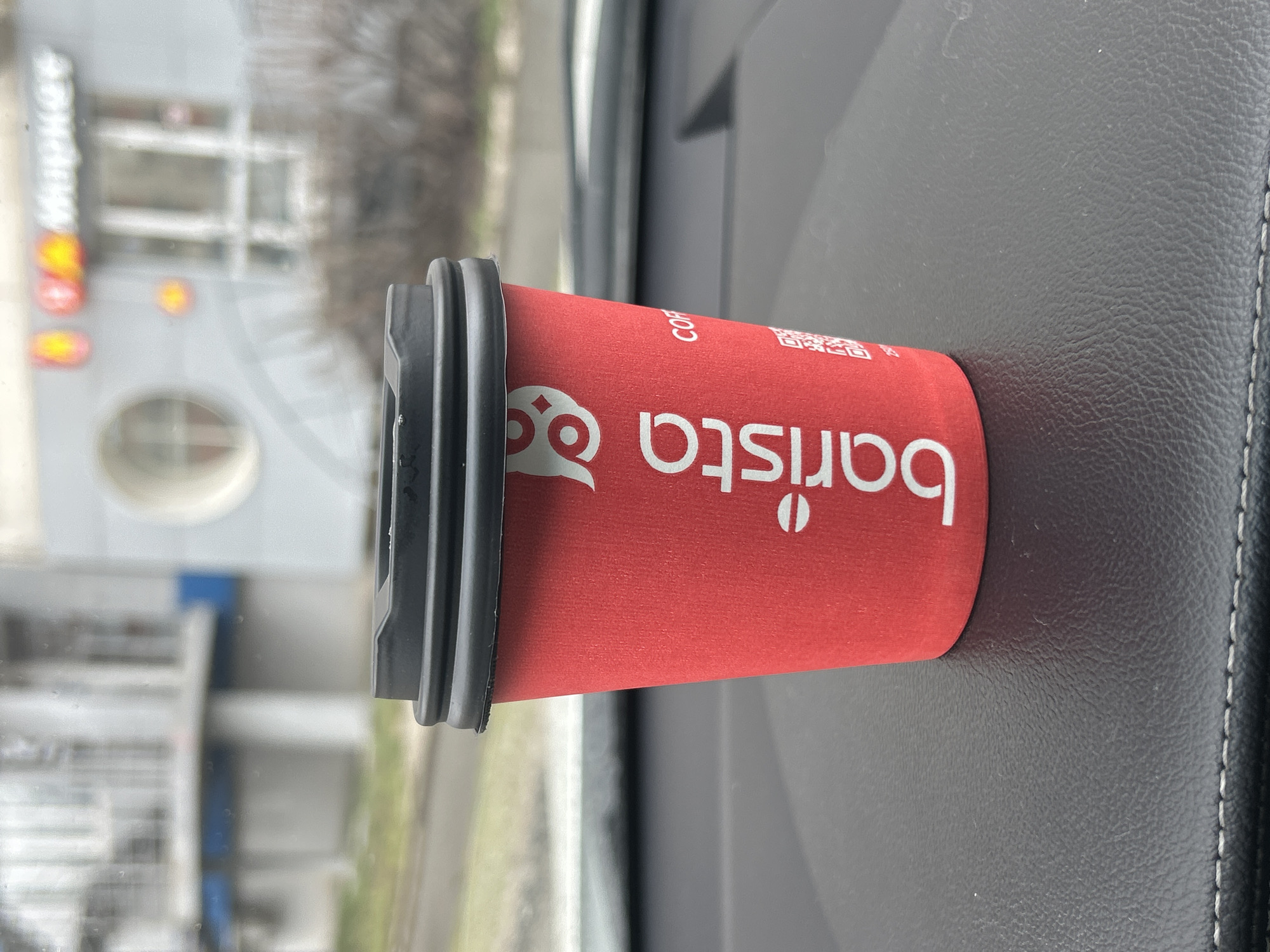 A red paper coffee cup with a black plastic lid is prominently displayed, resting on a dark, textured surface, likely a car dashboard or console, indicated by visible stitching on the right. The cup features the brand name "barista" in white script, along with a stylized white owl-like logo and the letters "COF" and a QR code, all against the red background.
The scene is set inside a vehicle, with the exterior visible through the windshield. The background is blurred but shows a building with reflective windows and several bright, yellow-orange lights that appear to be part of an illuminated sign. Partially legible text on the building sign indicates "MAгHИt" (Magnit), identifying it as a store belonging to the well-known Russian retail chain. This detail, combined with the provided location context, suggests the image was captured in Izhevsk, Russia.
The lighting appears to be daytime, possibly overcast, as there are no harsh shadows and the light is diffused. No people are visible, and the implied activity is simply someone consuming a beverage while in or shortly after being in a car, likely having just made a stop at the Magnit store.