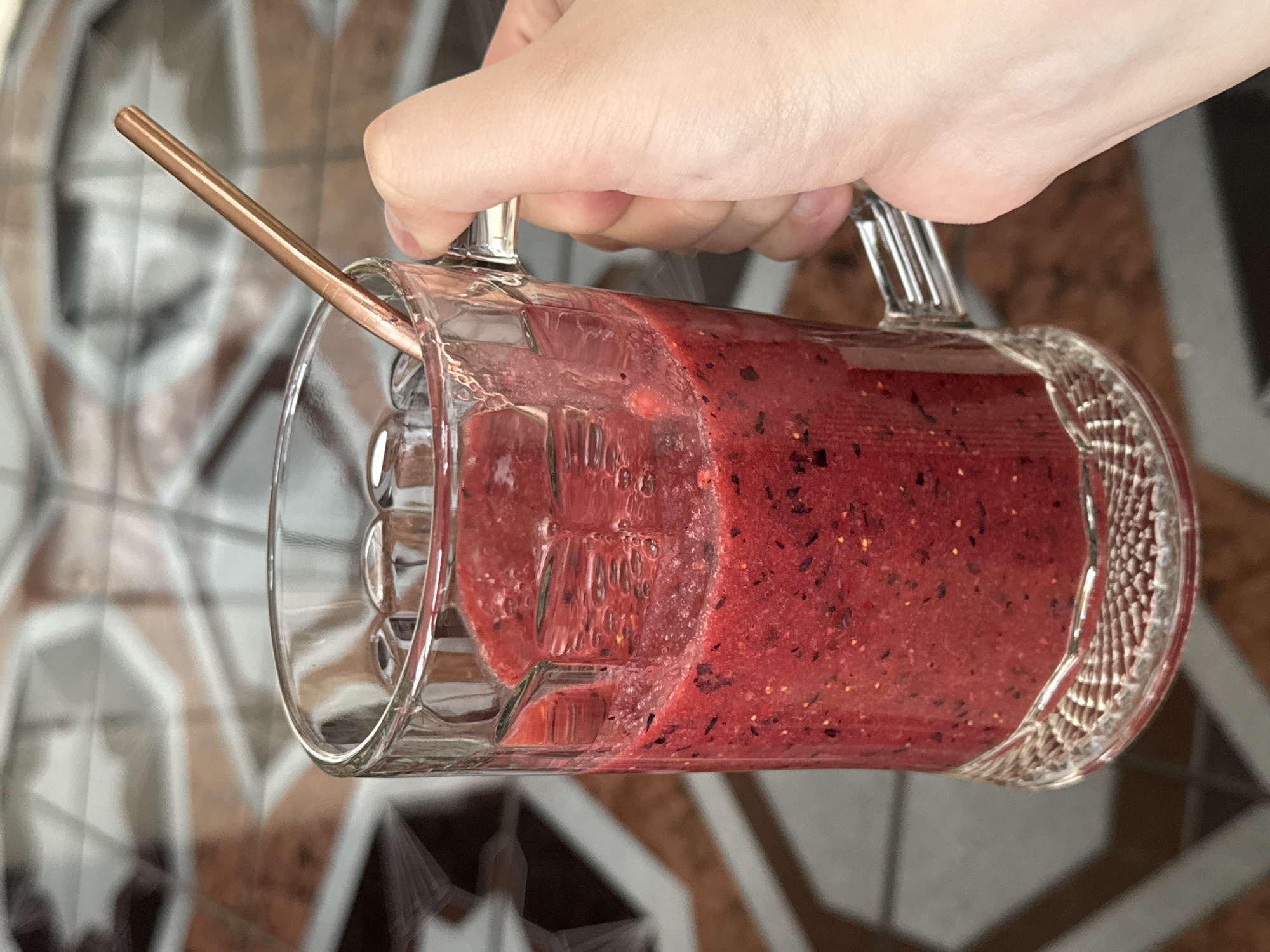 The image shows a close-up of a hand holding a clear glass mug filled with a red smoothie. A rose-gold metal straw is inserted into the smoothie. The mug has a handle and is made of thick, textured glass, with a decorative patterned rim. The smoothie itself appears to be made with berries, as evidenced by the small dark specks suspended within the red liquid. Bubbles are visible on the inside surface of the mug, suggesting the smoothie is fresh. The background is blurred, but appears to be an indoor setting with a tiled floor featuring a geometric pattern of brown, white, and grey tiles. The lighting suggests it is daytime. The context provided indicates the location is Moscow, Russia. There is no visible text in the image. The image focuses on the smoothie and the mug, with the hand and straw providing context for its use. The overall impression is one of refreshment and perhaps a healthy lifestyle.