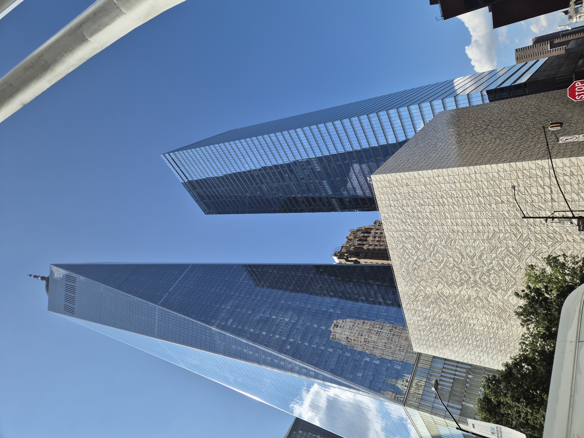 The image depicts the World Trade Center complex in New York City under a clear blue sky. Dominating the foreground is the distinctive facade of the 9/11 Memorial Museum, a large, angular building with a textured surface. To the left, the iconic One World Trade Center, a sleek glass skyscraper, rises dramatically, its reflective surface mirroring the sky and surrounding structures. To the right, another modern glass skyscraper, 3 World Trade Center, stands tall. In the background, other buildings of the complex are visible, including a gothic-style brick building, adding a historical contrast to the contemporary architecture. Trees and a white van are also present in the lower part of the frame, indicating an urban street-level perspective. A traffic light and a "STOP" sign are visible on the right side of the image. A banner reading "9/11 MEMORIAL MUSEUM" is affixed to a lamppost on the left.