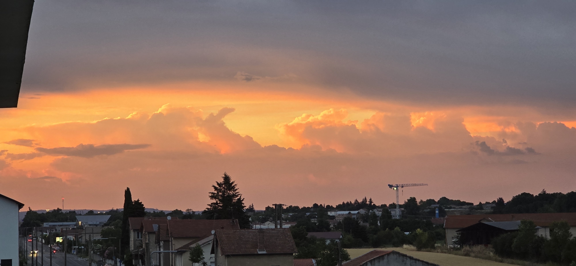 The image depicts a suburban landscape at dusk. A vibrant orange and pink sunset dominates the sky, casting a warm glow on the clouds. In the foreground, the roofs of several houses are visible, some with red tiles. A tall, dark green evergreen tree stands prominently on the left side of the frame, its silhouette sharp against the colorful sky. To the right of the tree, a construction crane is visible in the distance, its structure stark against the horizon. The rest of the scene is a blur of trees, buildings, and lights, suggesting a residential area. The overall impression is one of a peaceful evening in a quiet town.