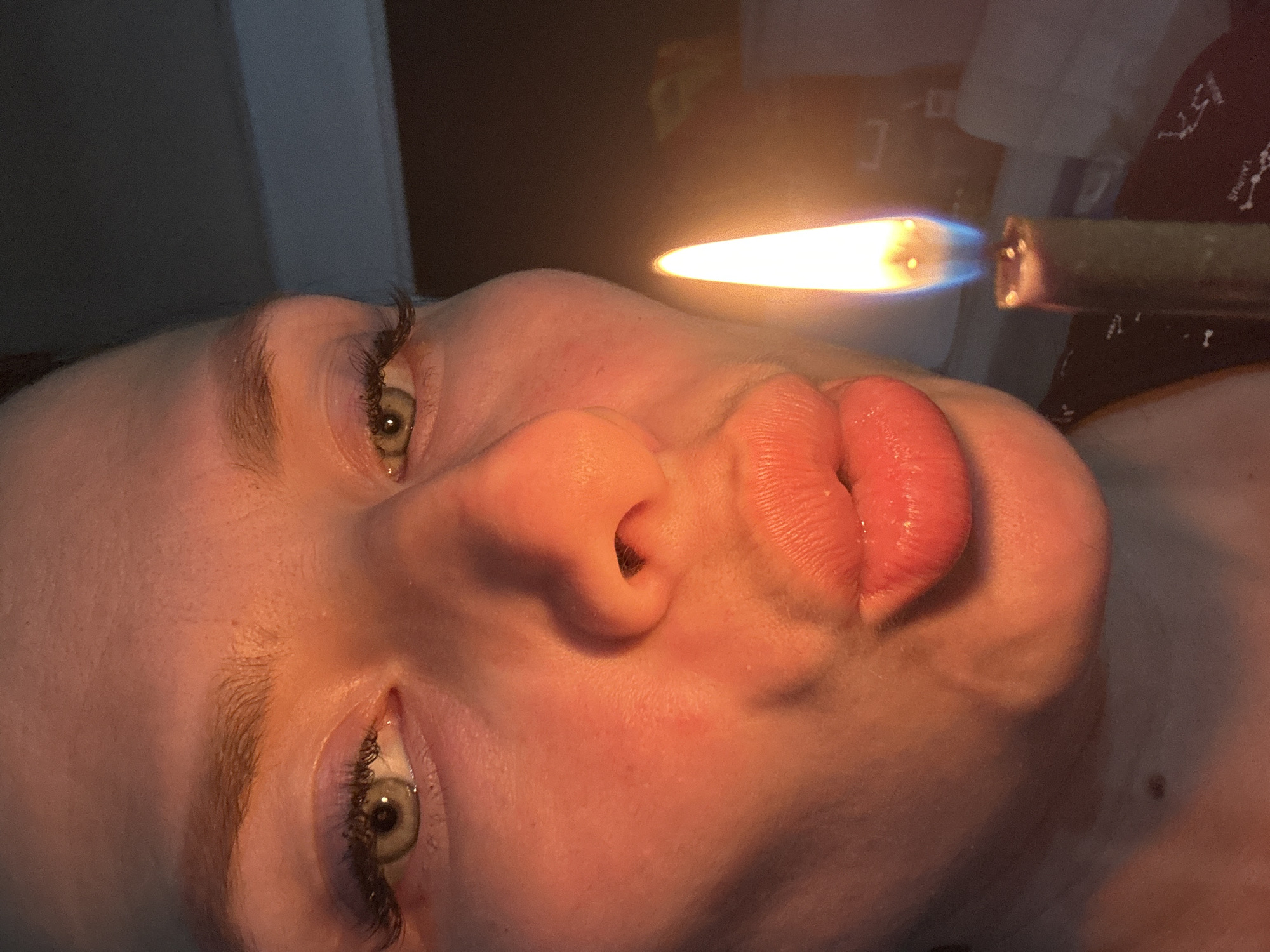 A close-up, angled view of a person's face, featuring green eyes and full lips. A flame from what appears to be a glass pipe is positioned near the face. The background contains a dark surface with fabric. The exact location is not discernible from the image, nor is the context to link it to Rusalevka, Russia.