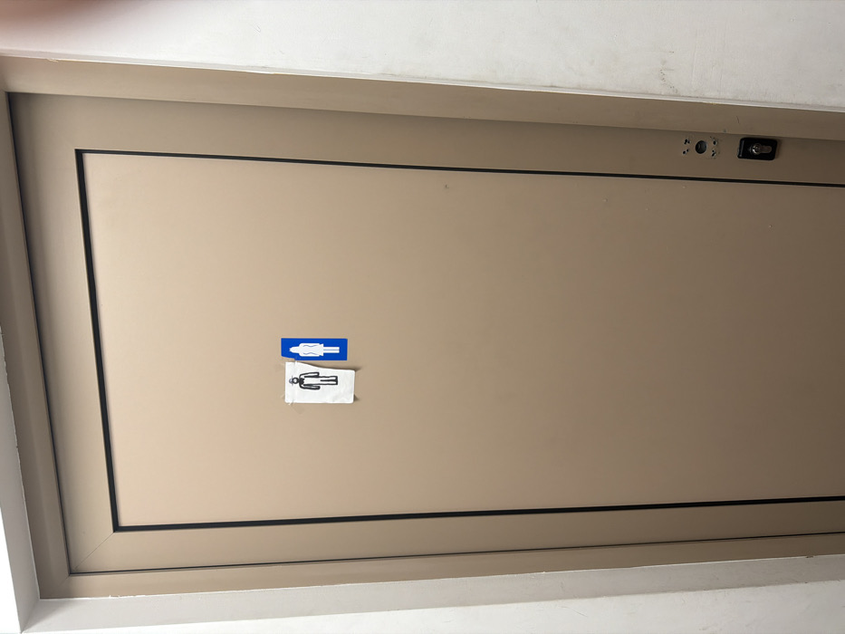 A closed, light-brown colored door, framed by dark trim, is depicted in an indoor environment in Antimovo, Bulgaria. Two restroom indicator signs are affixed to its surface, slightly right of the vertical center. The left sign, a black silhouette of a male figure with a beard and hard hat on a white, roughly cut piece of paper, is secured with visible adhesive tape. Immediately to its right is a blue rectangular sign with a white silhouette of a female figure. A keyhole and door handle plate are visible on the lower right side of the door. Portions of white plastered walls are present on both the left and right sides of the door frame. No individuals are present, and no actions or interactions are occurring.