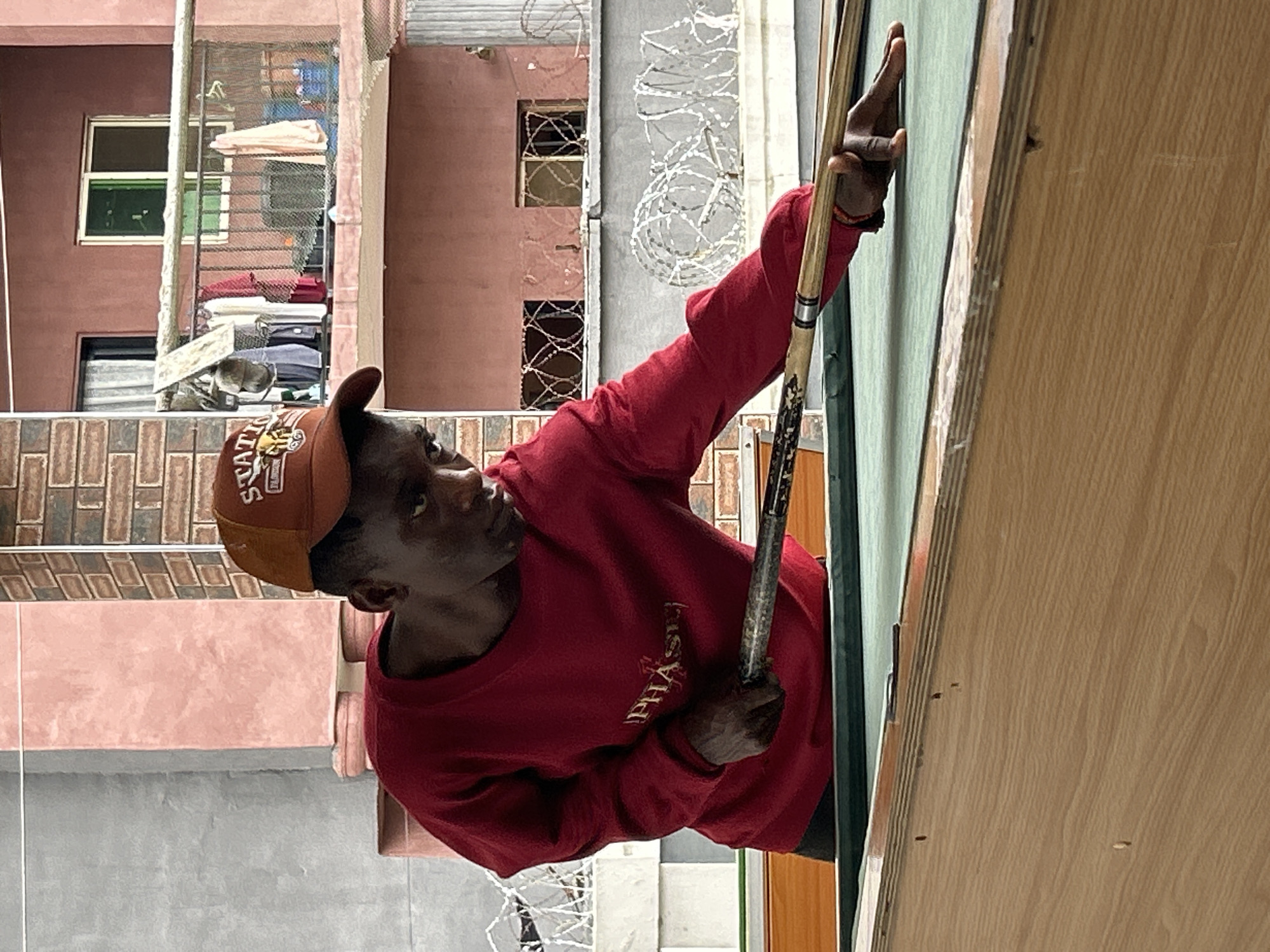 A young Black male wearing a red sweater and an orange baseball cap is playing pool. He is standing on the left side of a green pool table, with his right hand on the cue stick and his left hand supporting it on the table. He is looking down at the cue ball, focused on his shot. The background shows a building with a balcony where laundry is hanging. The image appears to be taken outdoors. The scene is set in the city of Ihiagwa, Nigeria.