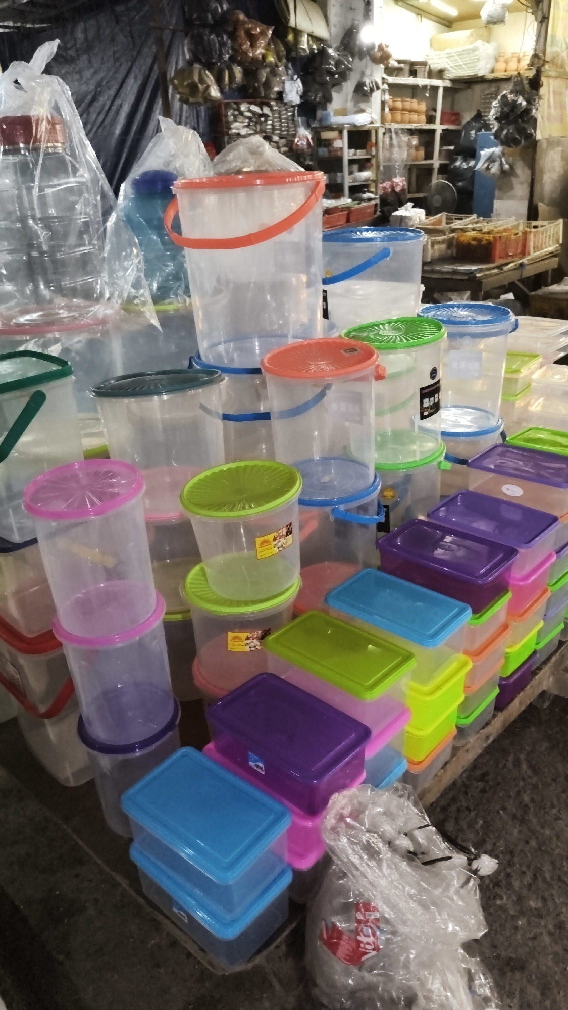 This image depicts a retail stall displaying a variety of plastic containers. The stall appears to be located in an outdoor or semi-outdoor market setting, possibly in South Jakarta, Indonesia, given the context of the request. The foreground is dominated by stacks of clear plastic buckets and rectangular storage containers in various sizes and colors, including red, blue, green, purple, and pink. Some of the buckets have red or blue handles and lids. In the background, there are shelves stocked with more goods, including what appear to be pottery or ceramic items, and other miscellaneous products. A large clear plastic bottle is visible on the left side of the frame, wrapped in plastic. The overall impression is of a busy market stall with an abundance of household storage items for sale.