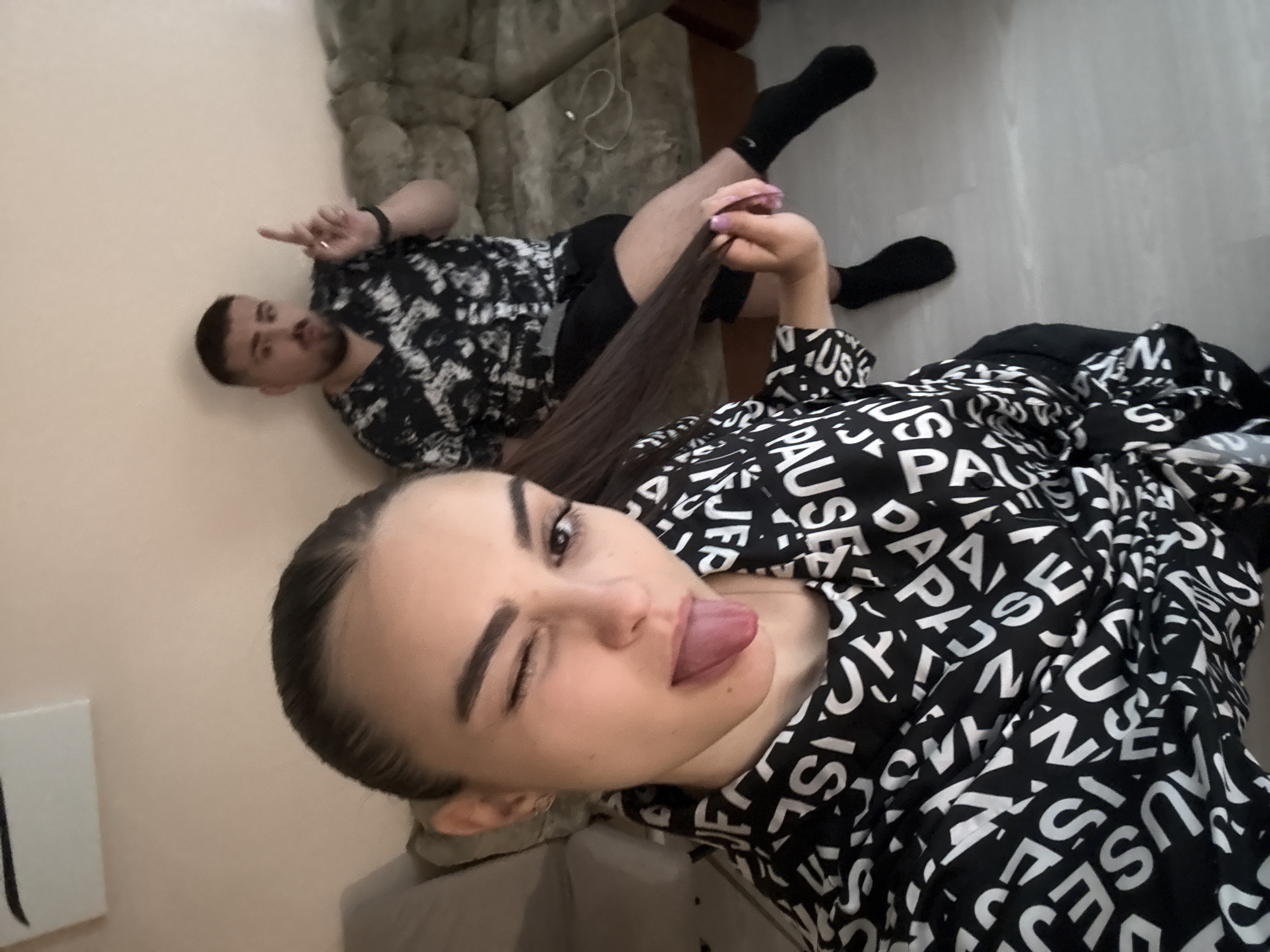 In an indoor setting, possibly a living room in Burgas, Bulgaria, a woman in the foreground takes a selfie. She is wearing a black and white patterned top and playfully sticks out her tongue while winking. Her dark hair is pulled back, and she is holding a section of it in her right hand. Behind her, a man is seated on a light-colored sofa, wearing a similar black and white patterned shirt and black shorts. He is looking towards the camera and has his right index finger raised, as if making a point. The room appears to be neutrally decorated with light-colored walls. A cable is visible on the sofa next to the man. The floor is a light grey laminate.
