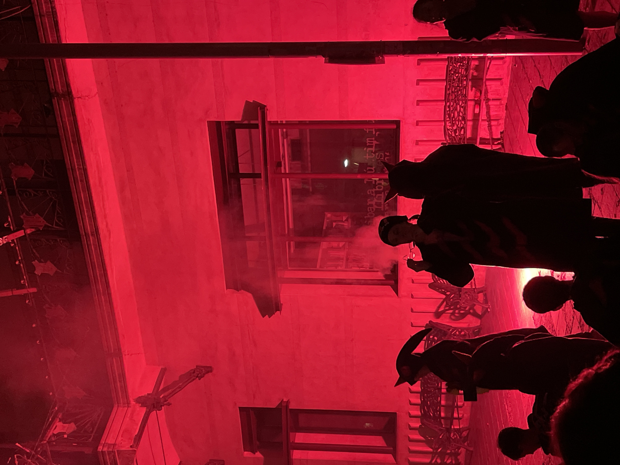 A group of people in silhouette are gathered in front of a building illuminated by red light. The building appears to be an older structure with architectural details visible, such as windows and decorative elements. Some of the people are wearing costumes, including one person dressed as the grim reaper and another with devil horns. The red lighting creates a dramatic and somewhat eerie atmosphere. In the background, there are benches and what appear to be decorative wrought iron elements. The scene suggests an outdoor event or performance, possibly a festival or theatrical production. The location is identified as Canet de Mar, Spain.