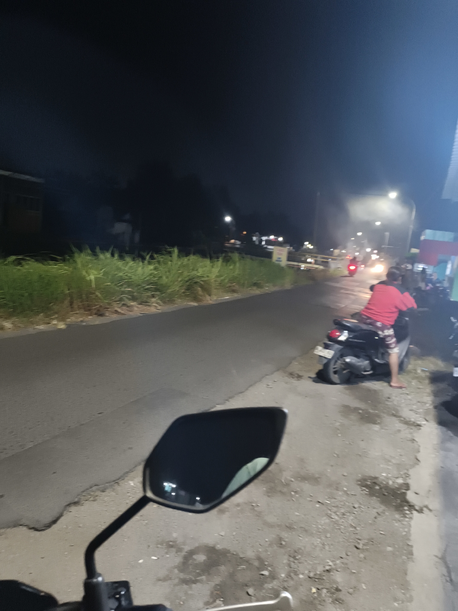 A nighttime outdoor shot captures a street scene in Sidorejo, Indonesia, with a focus on a person on a scooter. In the foreground, a black scooter handlebar and mirror are visible, suggesting the viewer is on another scooter. A person wearing a red shirt and patterned shorts is seated on a dark scooter on the right side of the road, facing away from the camera and towards the street. The scooter's license plate is partially obscured. The road is paved and appears wet, reflecting the light from nearby streetlights. On the left side of the road, a thick growth of green grass lines the roadside. Beyond the grass, a building with a dark facade is visible. In the background, other scooters and streetlights illuminate the scene, with a red scooter moving away from the viewer further down the road. The sky is dark, indicating it is nighttime.