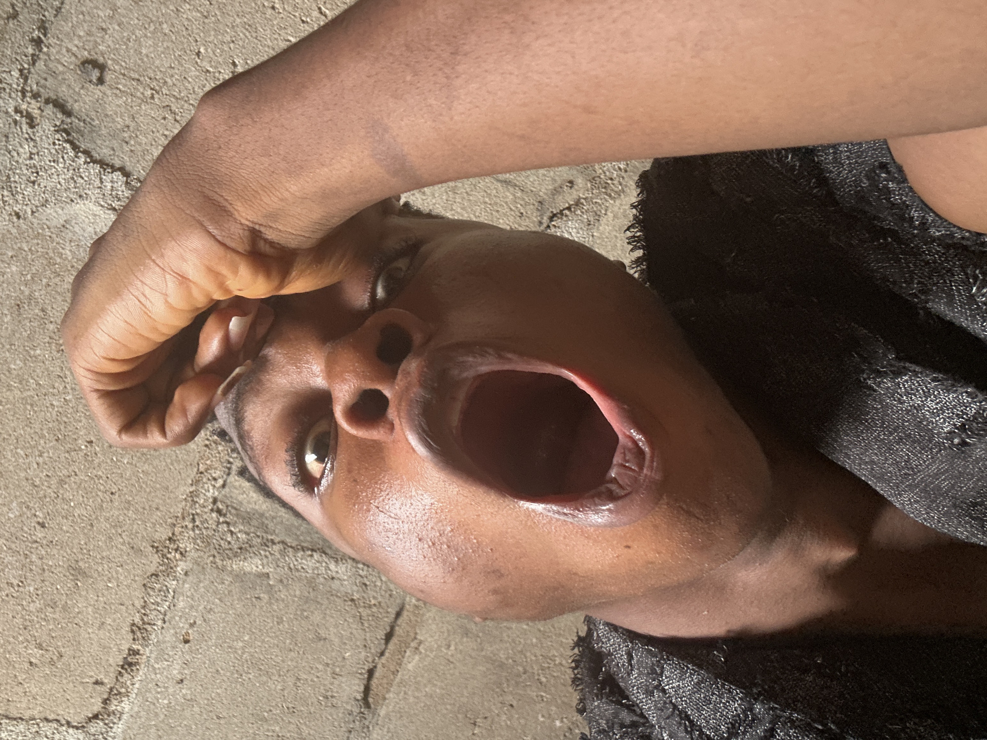 In Ikorodu, Nigeria, a person with dark skin is captured in a close-up, low-angle selfie against a textured, light-brown concrete wall. The individual's left hand is raised, with fingers pressed against their forehead. Their mouth is wide open in a yawn or shout, revealing their teeth and the inside of their mouth. They are wearing a dark, textured garment. The lighting suggests an outdoor setting, possibly during the day, with harsh shadows indicating direct sunlight.