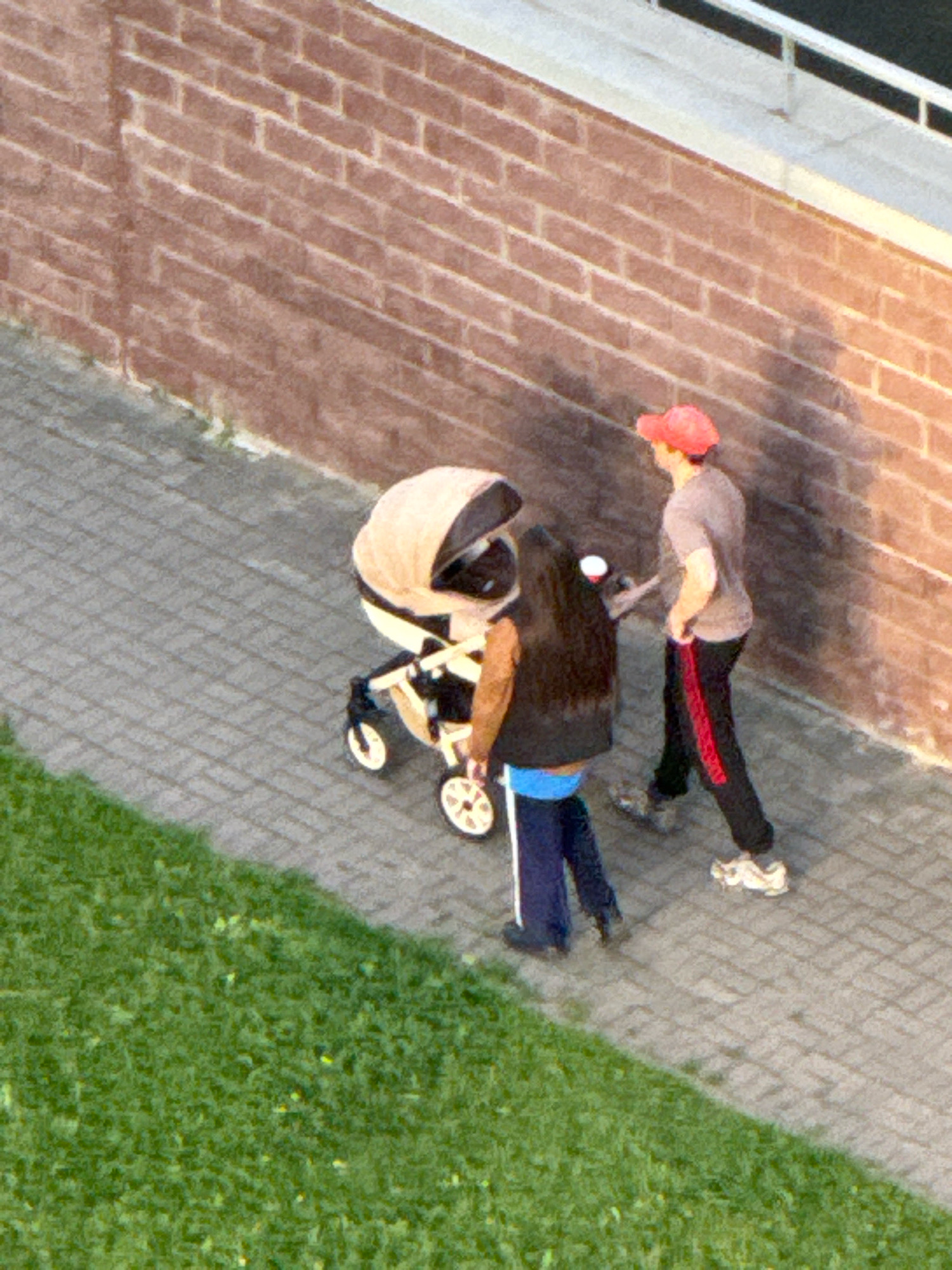 Two people are walking on a paved pathway next to a grassy area in front of a brick wall. The person on the right is wearing a red baseball cap, a grey t-shirt, and black pants with a red stripe down the side. They are pushing a baby stroller, which is tan and black. The person on the left is wearing a black vest over a blue shirt and dark pants, and is walking alongside the stroller. The paved pathway is made of interlocking grey bricks. The brick wall is made of reddish-brown bricks laid in a running bond pattern. The grassy area is green and appears to be a lawn.