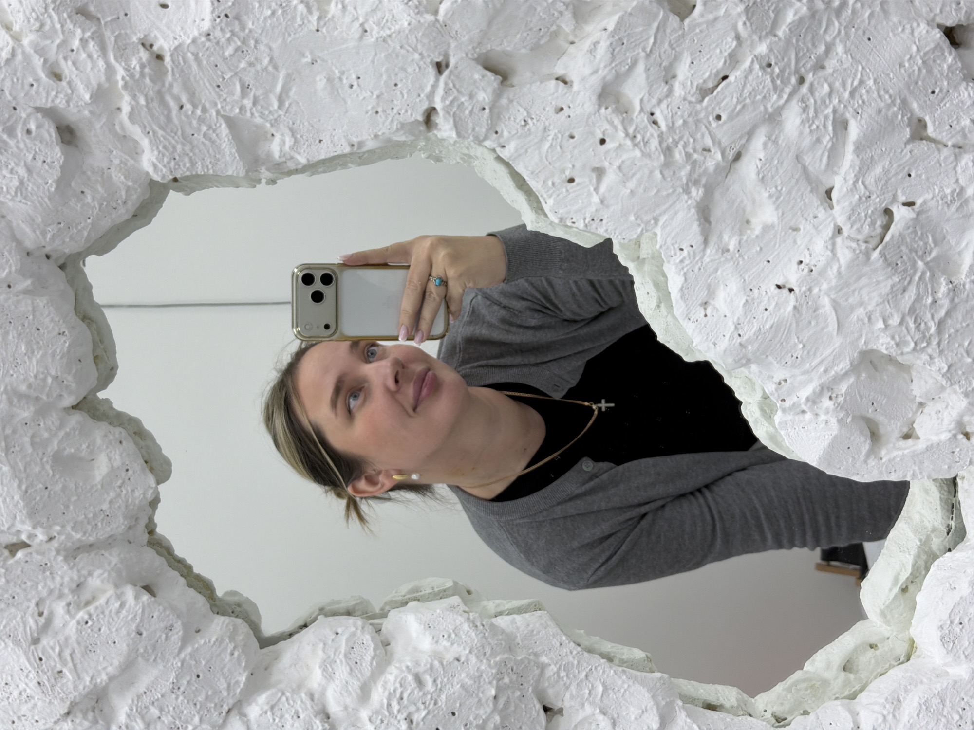 A woman is captured in a mirror selfie in an interior setting, identified as Moscow, Russia. The image focuses on the woman's reflection, which is framed by a highly textured, irregular white border around the mirror.
The woman, from the chest up, holds a gold-framed smartphone with a visible triple camera array to take the picture. She looks slightly upwards and to her left, displaying a calm and thoughtful expression. Her light-colored hair is partially pulled back, and she has light-colored eyes. She is dressed in a black top underneath a grey cardigan. Accessories include a gold chain necklace with a small cross pendant, a gold-toned earring on her right ear, and a ring with a blue stone on her right hand. Her fingernails are painted with a light pink polish.
The most prominent feature of the scene is the mirror's unique frame. It is made of a rough, sculptural white material, possibly plaster or concrete, with a highly uneven, organic surface featuring numerous indentations and raised areas, resembling an abstract, cave-like or rock formation. This distinctive frame creates an irregular opening for the mirror itself.
The background reflected in the mirror behind the woman is a plain white wall, suggesting a clean, brightly lit indoor space, such as a gallery or modern studio. The overall lighting is bright and diffused. There is no visible text within the image.