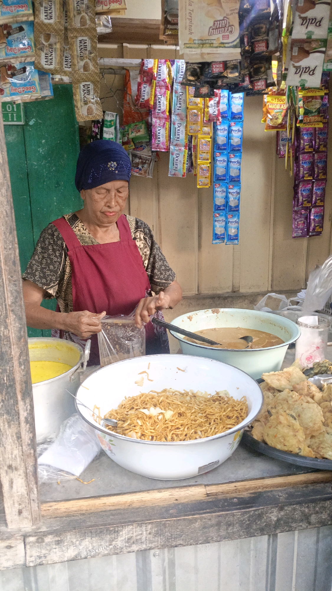 An elderly Indonesian woman, wearing a dark blue headscarf and a maroon apron over a patterned shirt, stands behind a wooden counter at a street food stall in Salatiga, Indonesia. She is in the process of packing food into a clear plastic bag, her hands holding the bag open. In front of her, a large white bowl overflows with stir-fried noodles. To her right, a light blue basin contains a light brown soup or curry, with a ladle resting in it. Further to the right, a black platter holds several pieces of fried food, possibly tempeh or fritters. A yellow pot is visible to the left of the woman. The stall's backdrop is a light-colored wooden wall adorned with various hanging packets of instant noodles, snacks, and personal care products, such as shampoo and fabric softener. The overall scene depicts a bustling street food vendor preparing to serve customers.