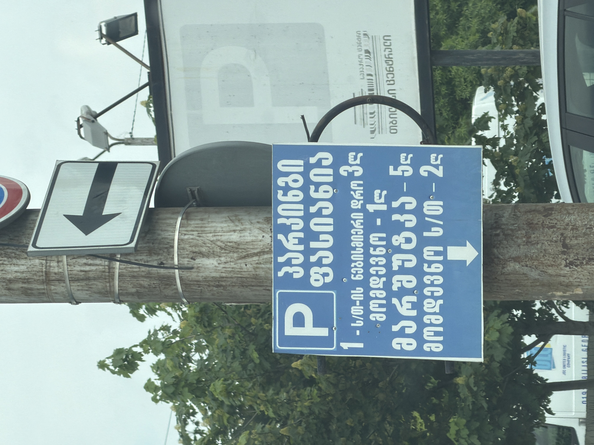 The image shows a parking sign in Tbilisi, Georgia. The sign is blue with white lettering and a white "P" indicating parking. The text on the sign, written in Georgian, specifies the parking costs: 3 GEL for the first hour, 1 GEL for each subsequent hour, 5 GEL for a marshrutka (a type of public transportation), and 2 GEL for each subsequent hour. An arrow on the sign points to the right. The sign is attached to a wooden pole along with other traffic signs, including a downward-pointing arrow sign. The environment includes trees and a glimpse of a building, possibly associated with JSC United Logistic Company, as seen on the bottom right of the image. A white vehicle is also partly visible.