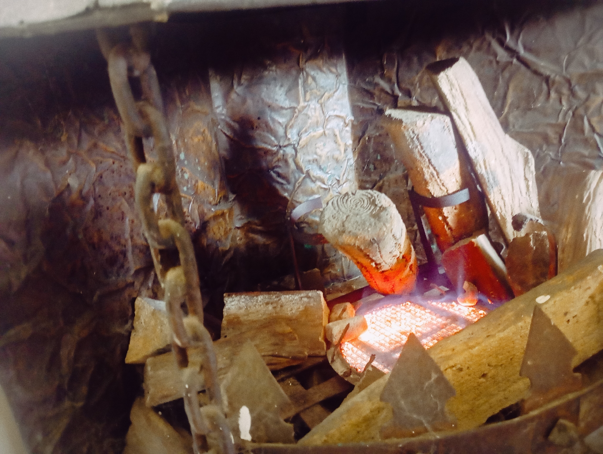 An upward-facing shot captures a detail of a simulated fireplace or heater, suggestive of a rustic or industrial aesthetic. In the foreground on the left, a thick, rusty metal chain hangs vertically, its links prominent. To the right of the chain, several irregularly shaped pieces, resembling ceramic logs or artificial wood, are arranged within what appears to be a firebox. These pieces vary in size and shape, some appearing charred or worn, with one featuring a swirl pattern on its top and another secured by a dark band.
At the bottom center and right, a bright, red-orange glowing heating element is visible, resembling an incandescent grate, indicating that the heater is active. This element casts a warm, fiery light on the surrounding artificial logs, giving the impression of burning embers. The background consists of a crumpled, textured surface, possibly metallic sheeting or foil, suggesting a rough, sooty interior wall of a fireplace. The overall scene conveys an artificial warmth and a decorative interpretation of a traditional fire.
No people or identifiable text are present. The image provides no specific time of day or weather cues beyond the implication of a need for warmth. Given the context of Buenos Aires, Argentina, this could be a functional heater or a decorative element in a home, restaurant, or public space, especially during cooler seasons.