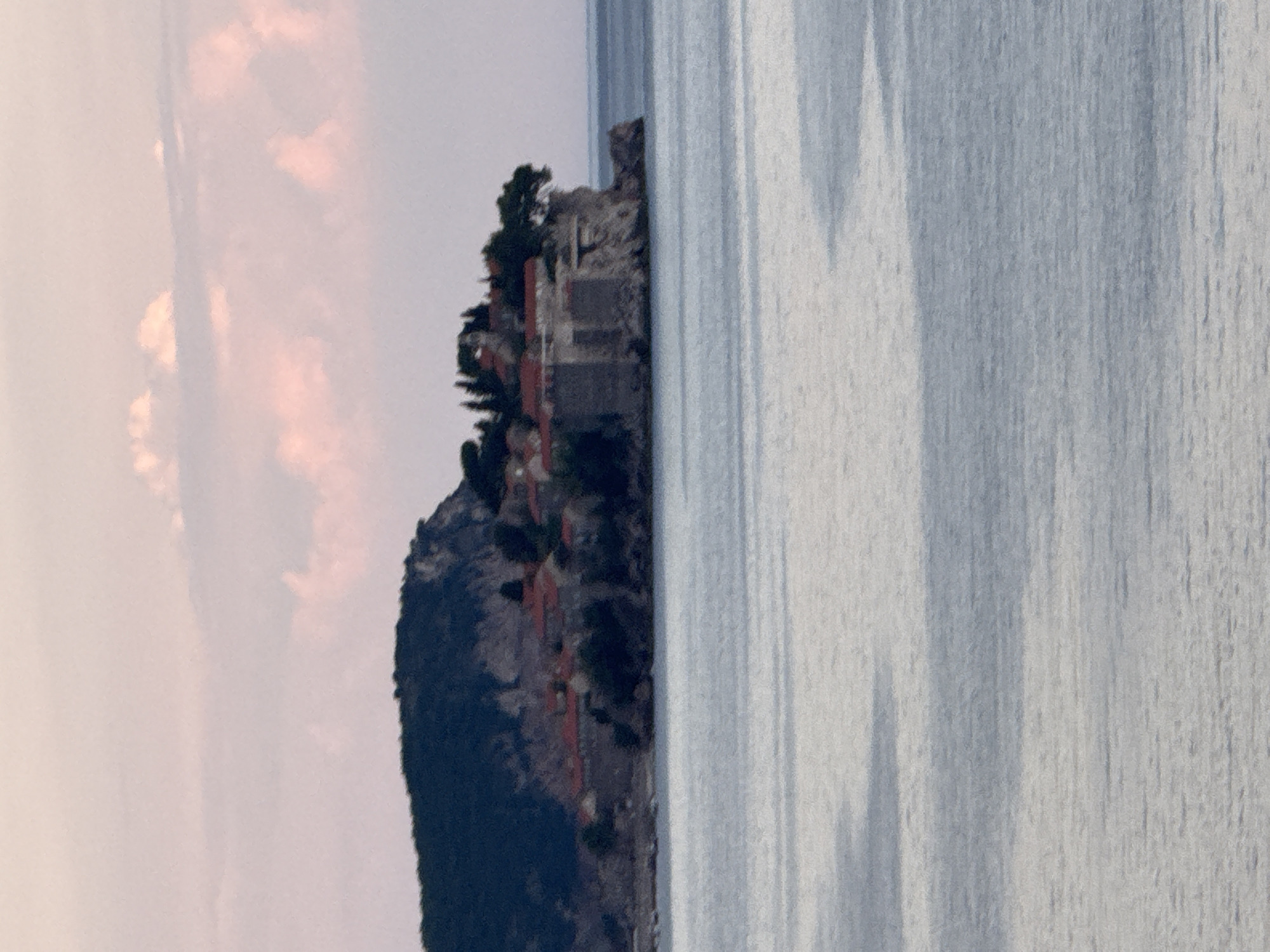 This media file shows a scenic view of a cliffside with buildings and vegetation overlooking the sea. The image is oriented vertically, with the cliff and buildings on the left and the sea on the right. The sky, visible in the upper left portion of the image, is a soft, muted color, suggesting either dawn or dusk, with some wispy clouds that appear to be tinged with pink and orange hues.
The cliff itself is rugged and covered in dark green vegetation, with several red-roofed buildings nestled into its side. The architecture of these buildings appears to be rustic, possibly stone or stucco, with some having multiple levels. There's a sense of a small settlement or a collection of dwellings built into the natural landscape. A larger, more imposing stone structure, possibly a fortification or an old building, is visible near the top of the cliff.
The sea on the right side of the image is calm and reflects the sky, creating a textured surface with variations in light and shadow. The reflections suggest a slightly overcast or hazy sky, but also possible sunlight creating streaks of lighter and darker blue/grey. The overall impression is one of tranquility and natural beauty, with a touch of historical or man-made presence integrated into the environment. The location context suggests this is Kamenovo, Montenegro, a place known for its coastline and picturesque villages. No text is visible in the image.